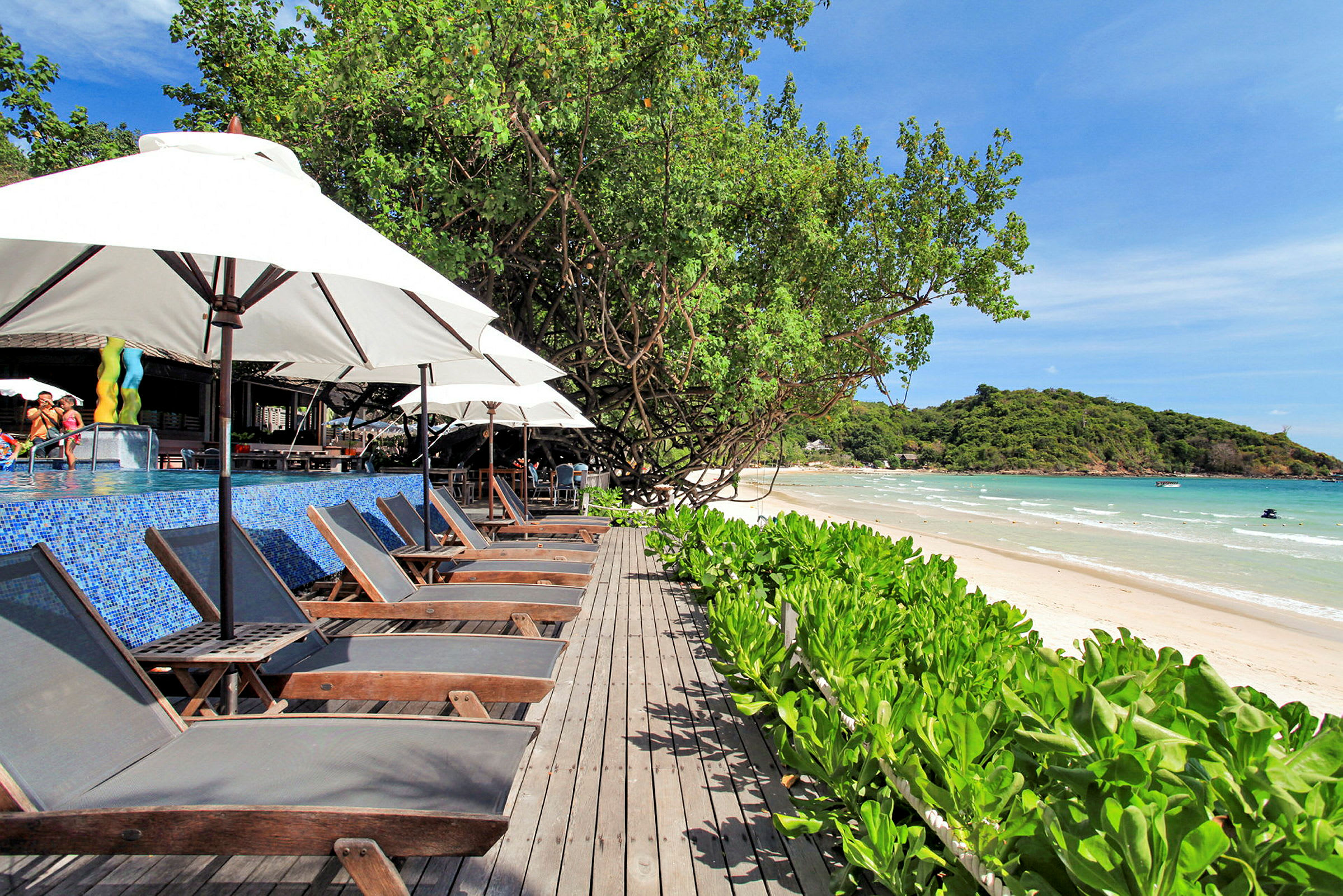 En strand med parasoller og solstoler langs en treterrasse, med utsikt over havet og vegetasjon. (AI-generert tekst)