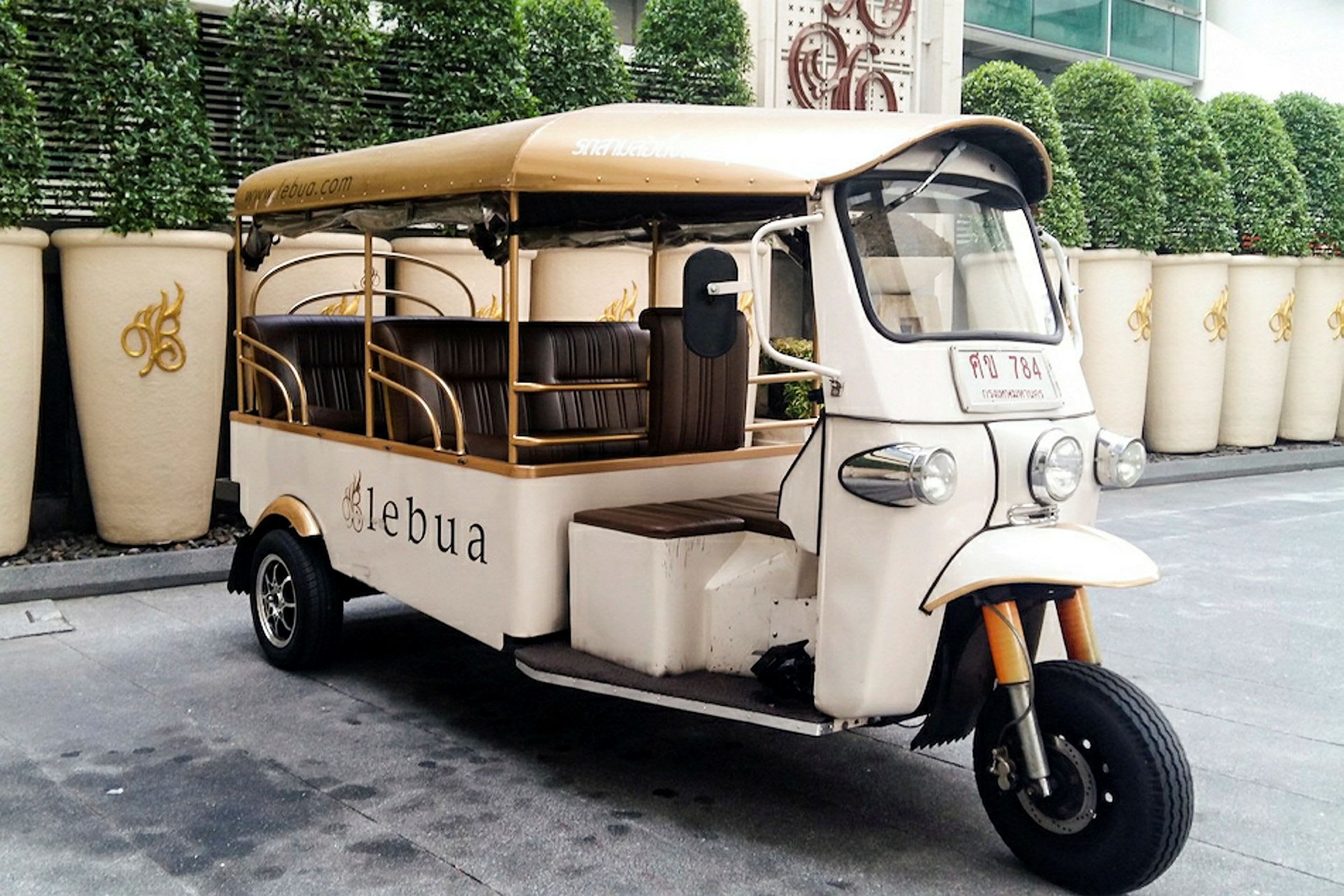 En hvit tuk-tuk med beige tak står parkert ved siden av en rad store hvite krukker og grønne planter i bakgrunnen. (AI-generert tekst)