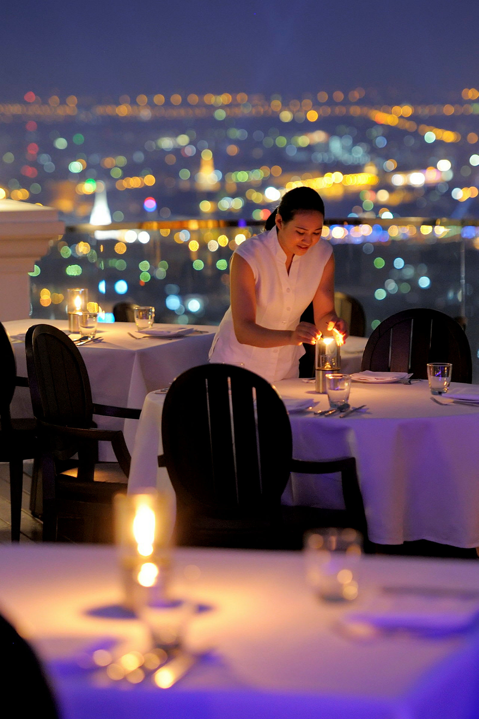 En person tenner lys på et dekket bord i en restaurant med utsikt over en bys silhuett. (AI-generert tekst)
