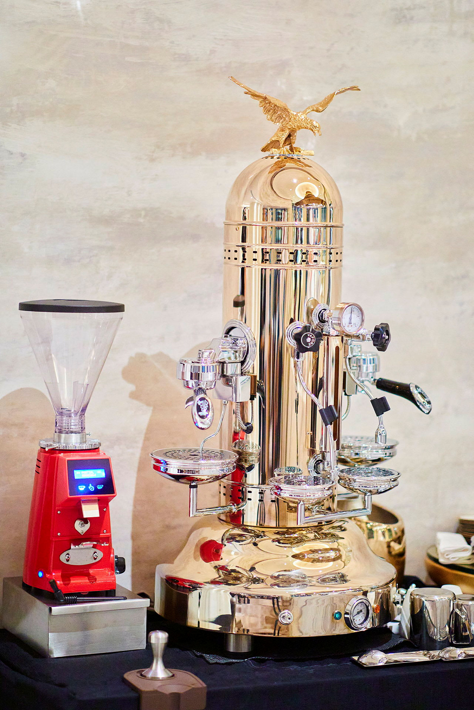 En elegant kaffemaskin i metallisk farge med flere bryggehoder og detaljer ved siden av en rød kaffekvern. (AI-generert tekst)