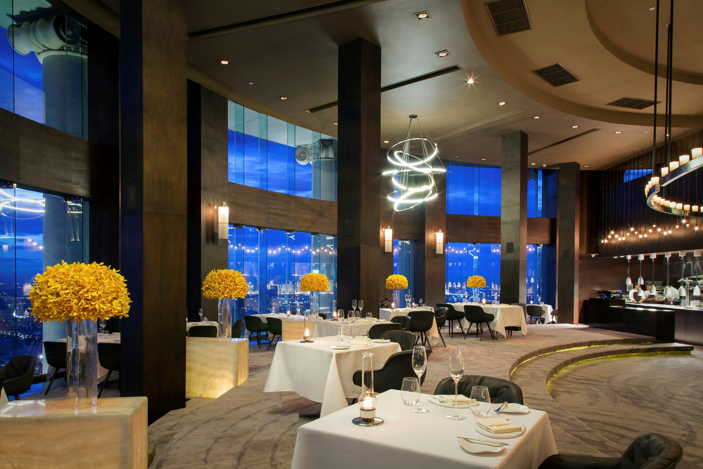En elegant restaurant med dekkede bord, moderne møbler og store vinduer mot en by. Gule blomsterdekorasjoner er plassert på pidestaller. (AI-generert tekst)