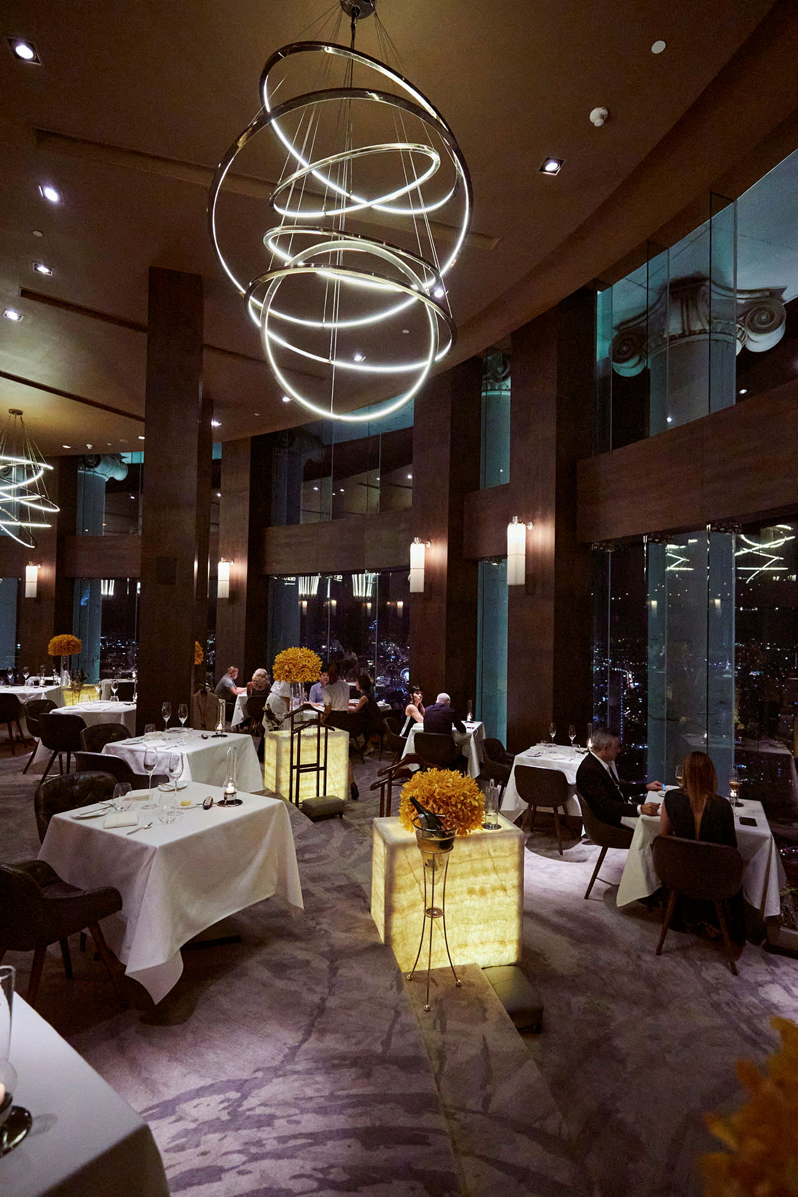 En luksuriøs restaurant med vinduer som viser bybilder om kvelden. I forgrunnen vises bord med borddekorasjoner. (AI-generert tekst)