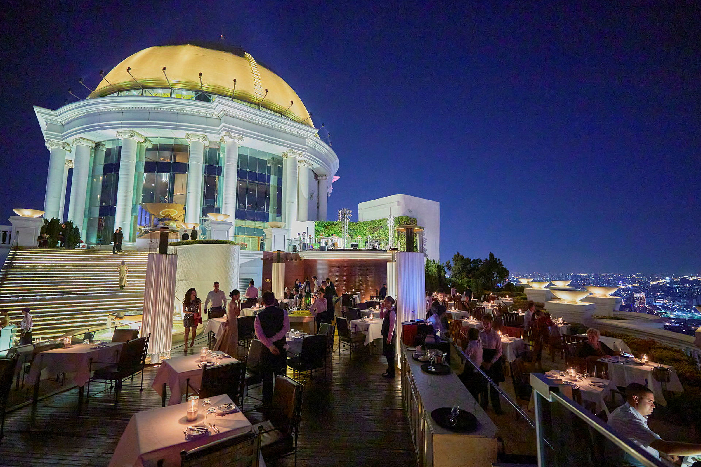 En restaurant på en takterrasse med utsikt over en opplyst by i skumringen. Bildet viser bord, gjester og servitører. (AI-generert tekst)