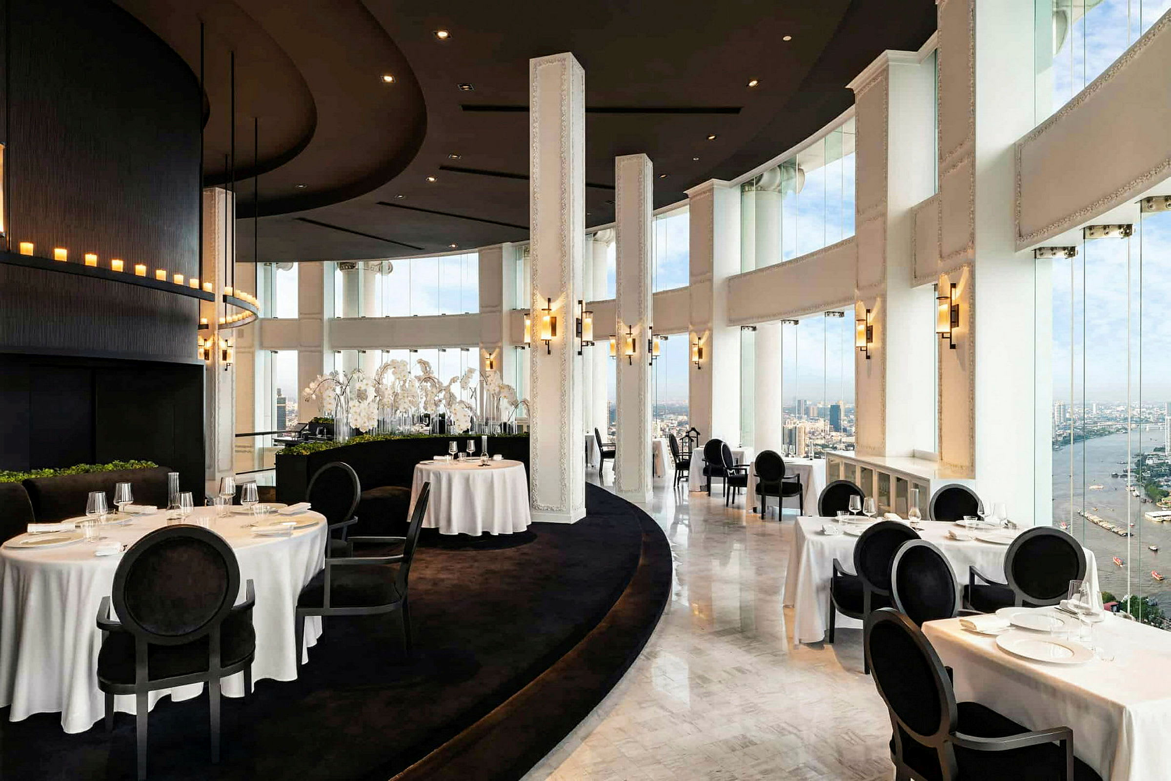 En elegant restaurant med store vinduer og hvit borddekking, som gir utsikt over byen og en elv. (AI-generert tekst)