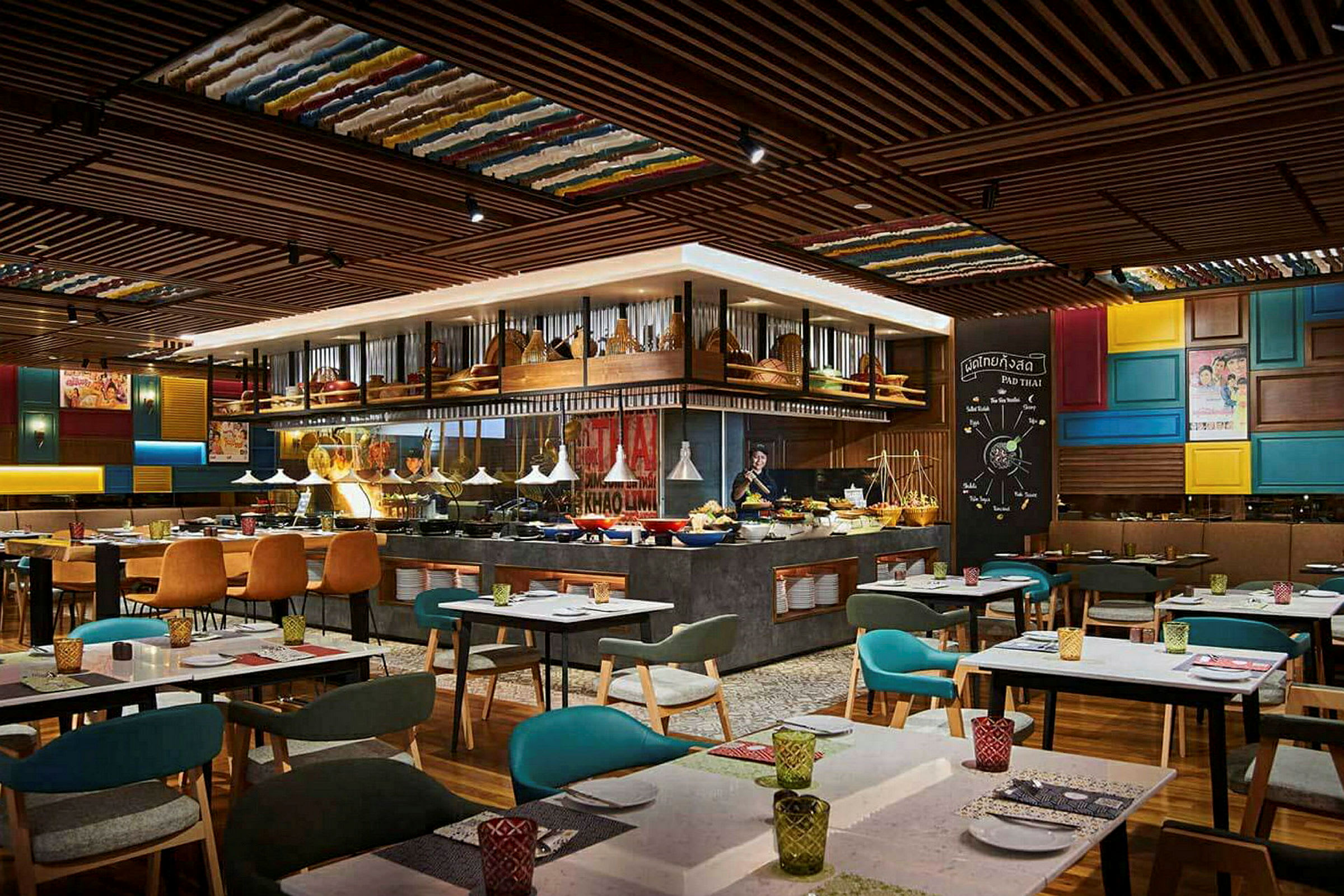 Restaurantinteriør med bord, stoler og en matbuffé i midten, omgitt av tretak og fargerike dekorative elementer. (AI-generert tekst)