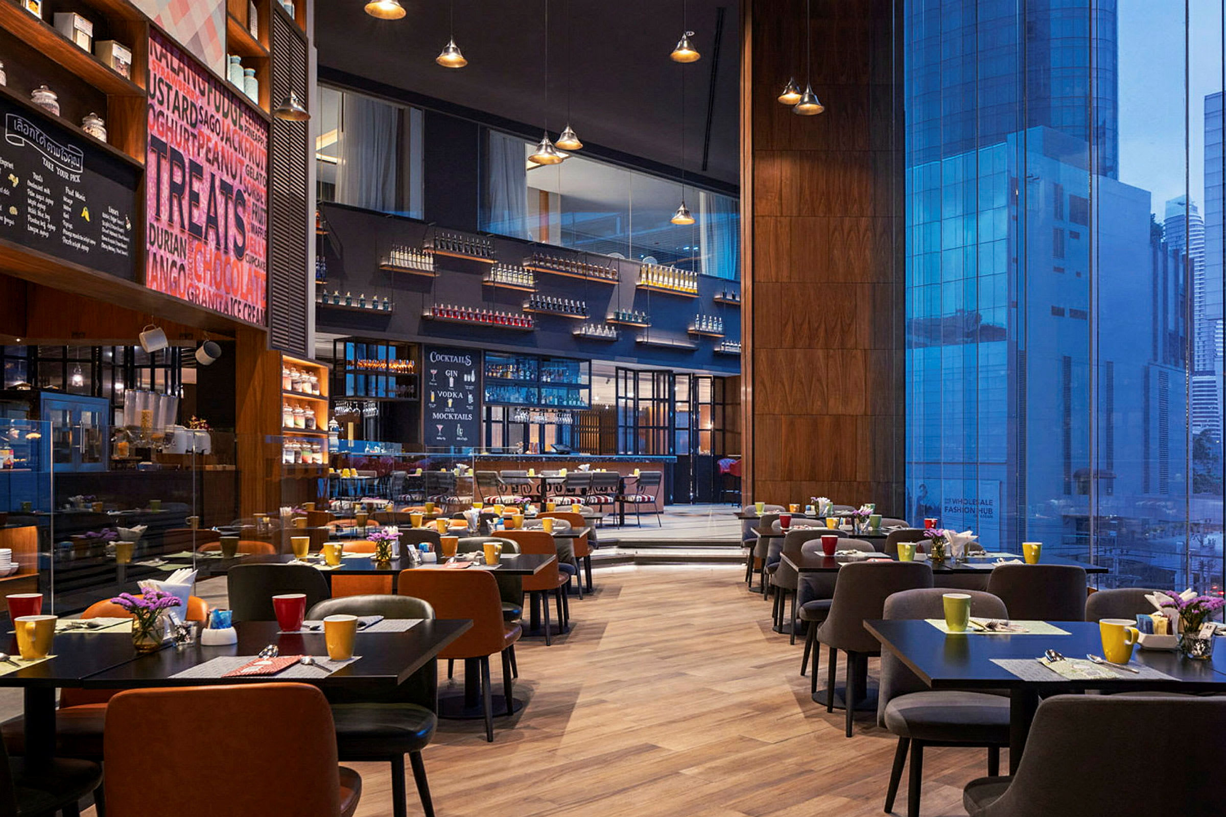 En romslig restaurant med moderne bord og stoler oppstilt, fargerike kopper og blomster på bordene og en stor glassvegg som viser utsikt over byens bygninger. (AI-generert tekst)