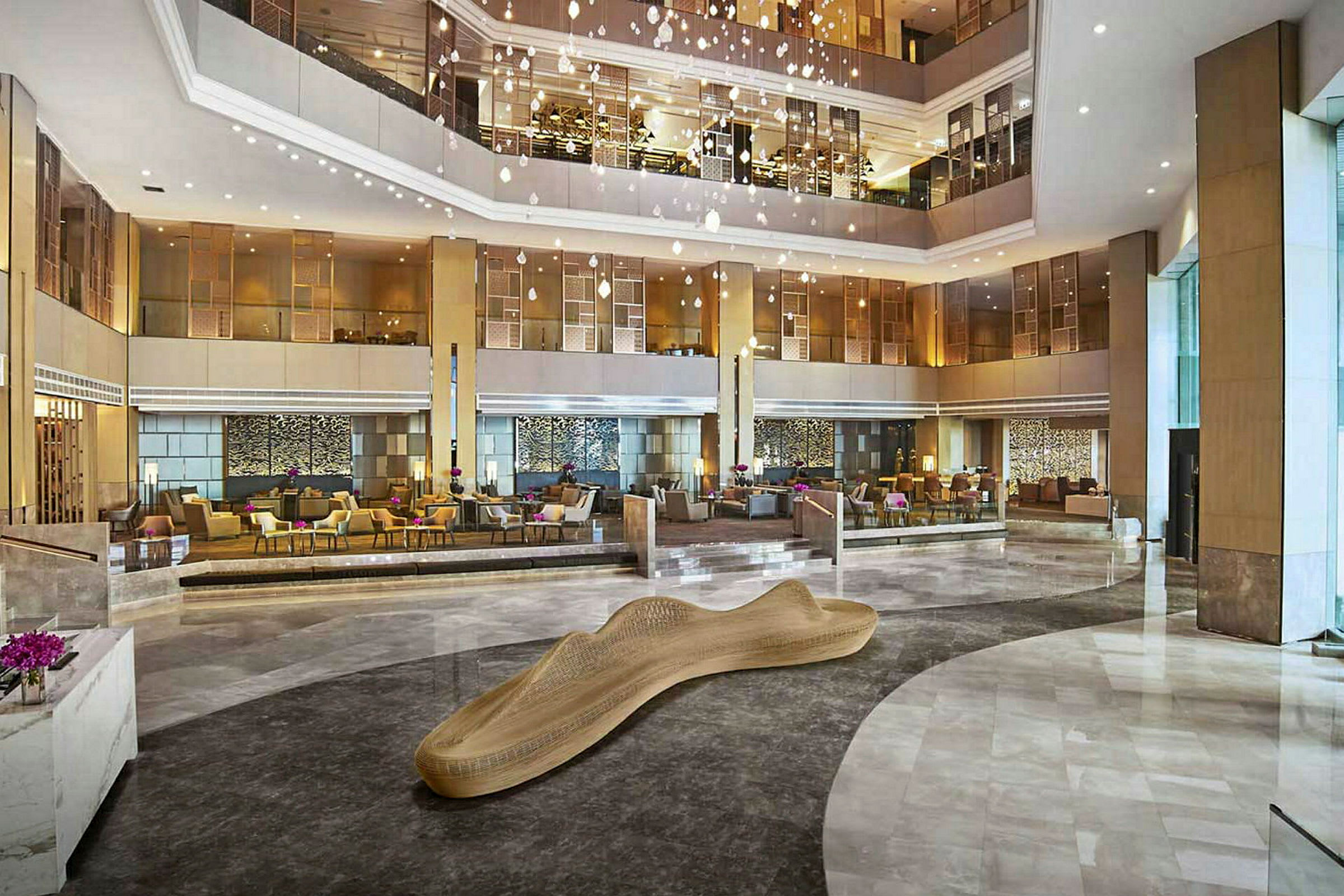 Et stort hotellobbyområde med moderne møbler og en dekorativ treskulptur i midten. Flere sittegrupper og elegant belysning. (AI-generert tekst)