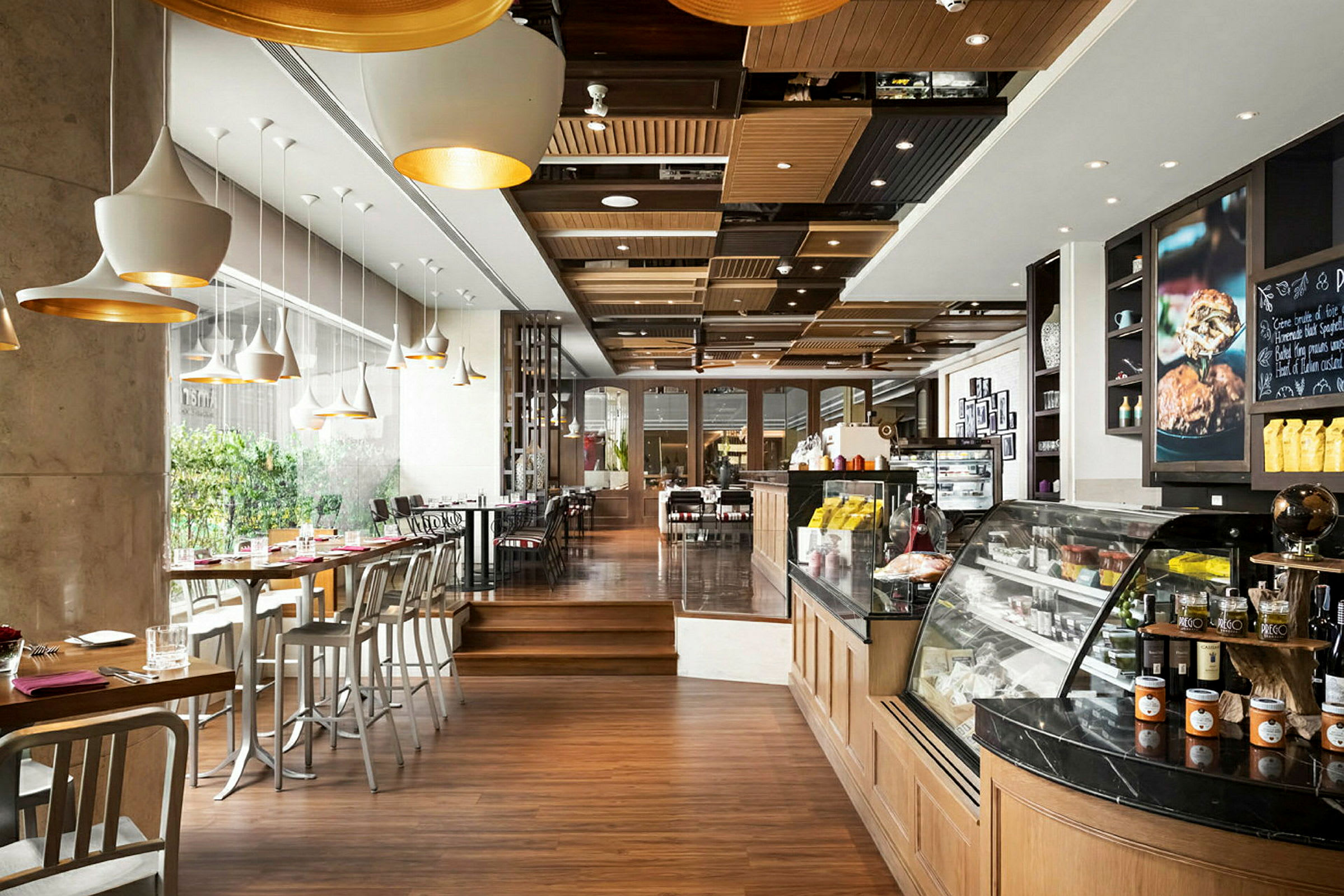 Et moderne kafé med tregulv, store vinduer og sitteplasser ved bord og barstoler. En opplyst disk med mat og drikke er på høyre side. (AI-generert tekst)