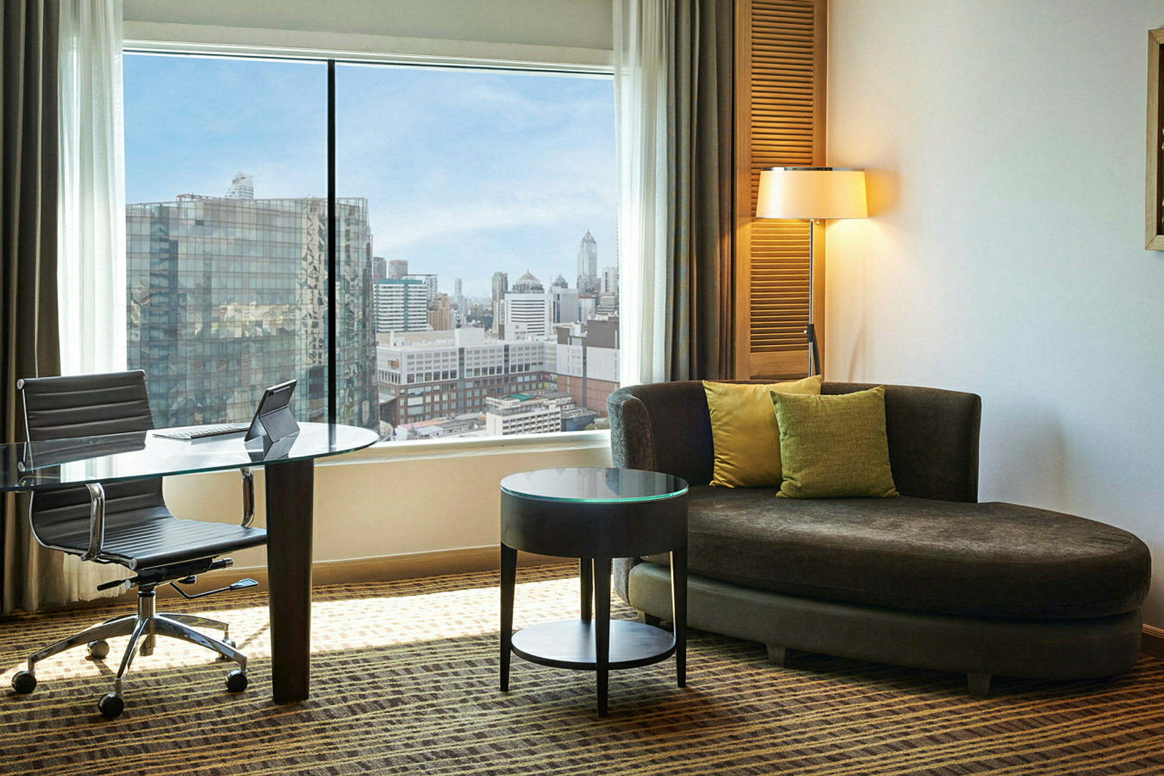 Et hotellrom med stor vindusutsikt over en by. Det finnes et glassbord og stol ved vinduet, samt en brun sofa med puter og et rundt sidebord. (AI-generert tekst)