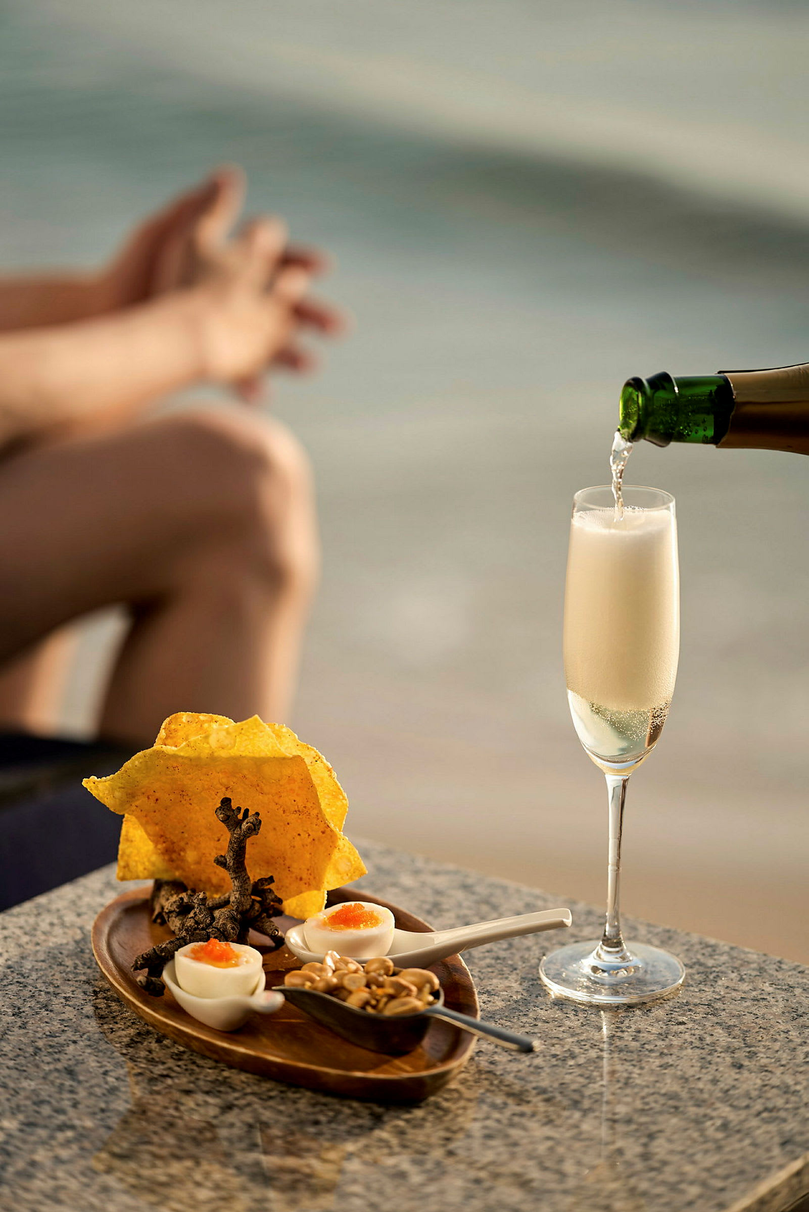 Et glass champagne blir skjenket på et bord med en liten servering av snacks foran en uklar bakgrunn av strand og hav. (AI-generert tekst)