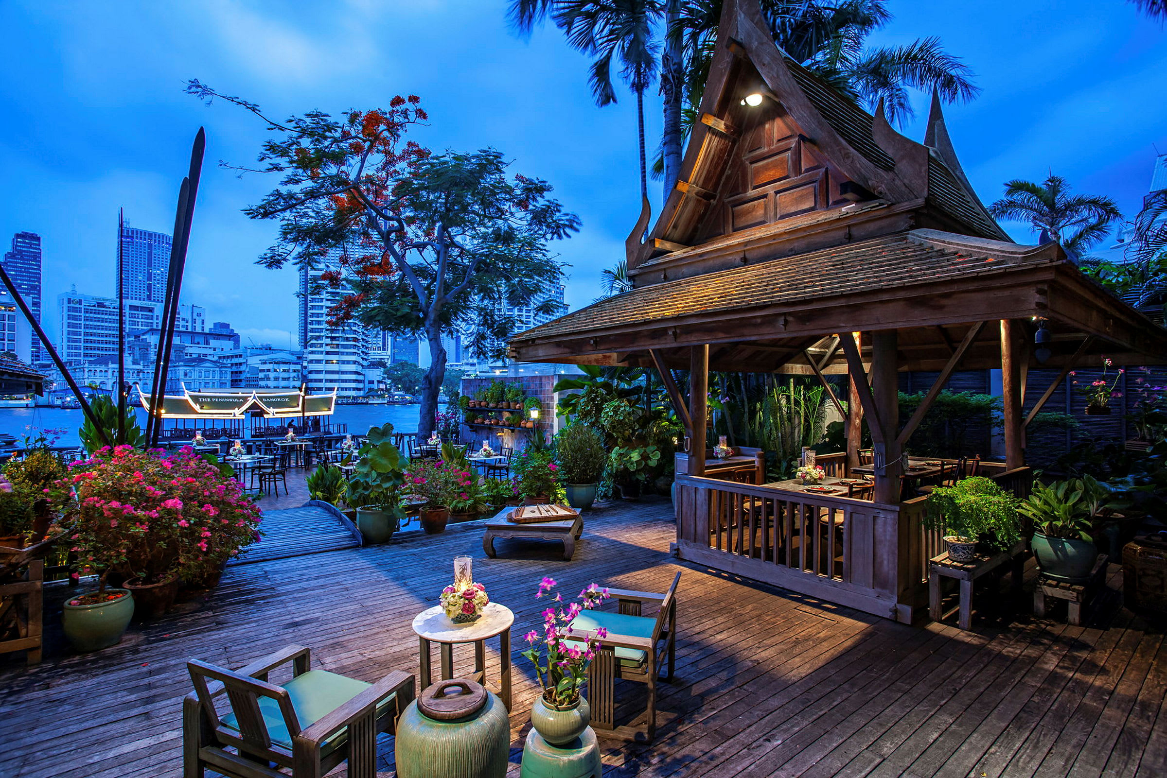 Et opplyst trehus med tradisjonell thailandsk design står på en terrasse omgitt av blomster og planter. I bakgrunnen sees en elv og byens silhuett ved skumringen. (AI-generert tekst)