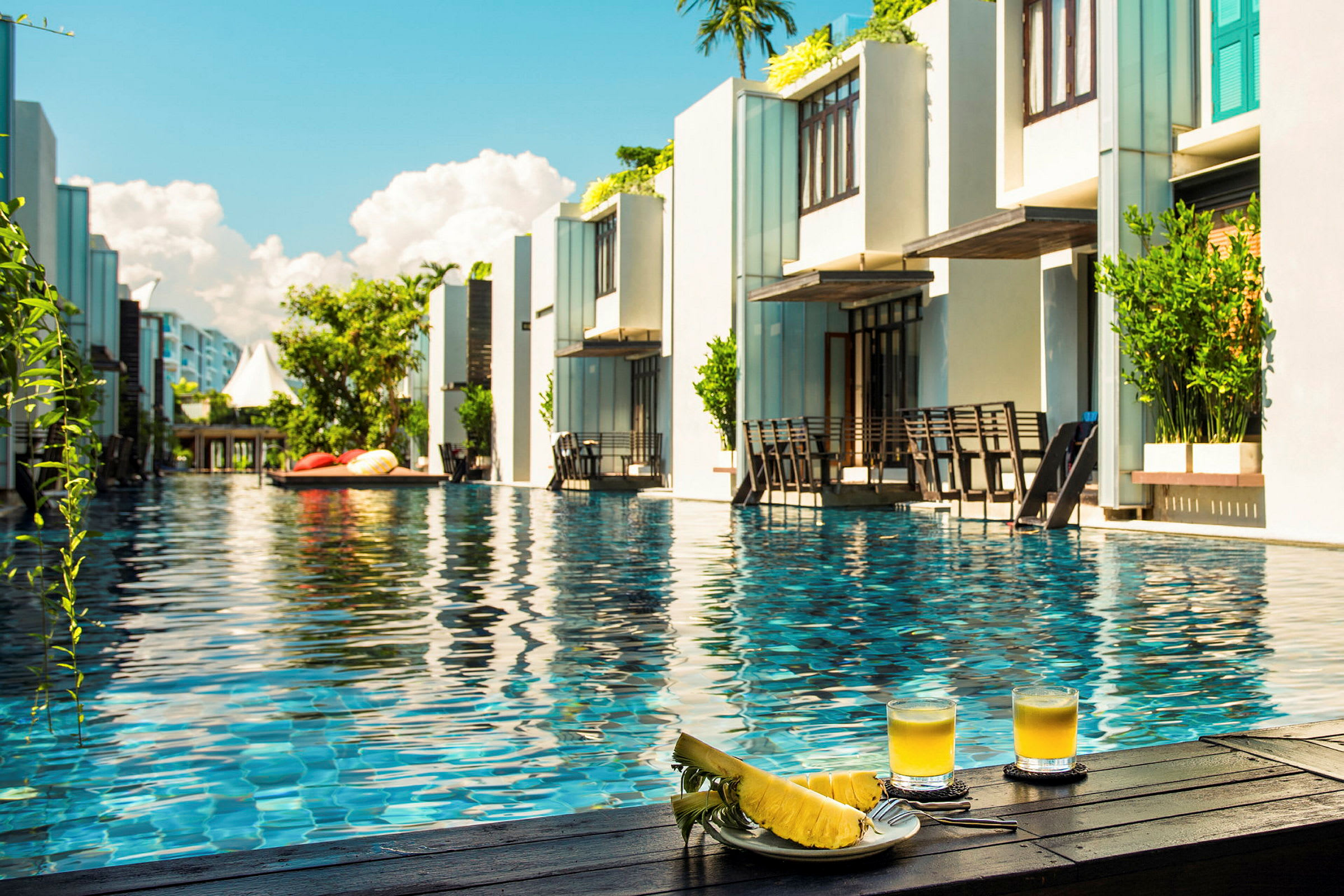 Resortbasseng omgitt av moderne bygninger med to glass juice og skivet ananas i forgrunnen. (AI-generert tekst)