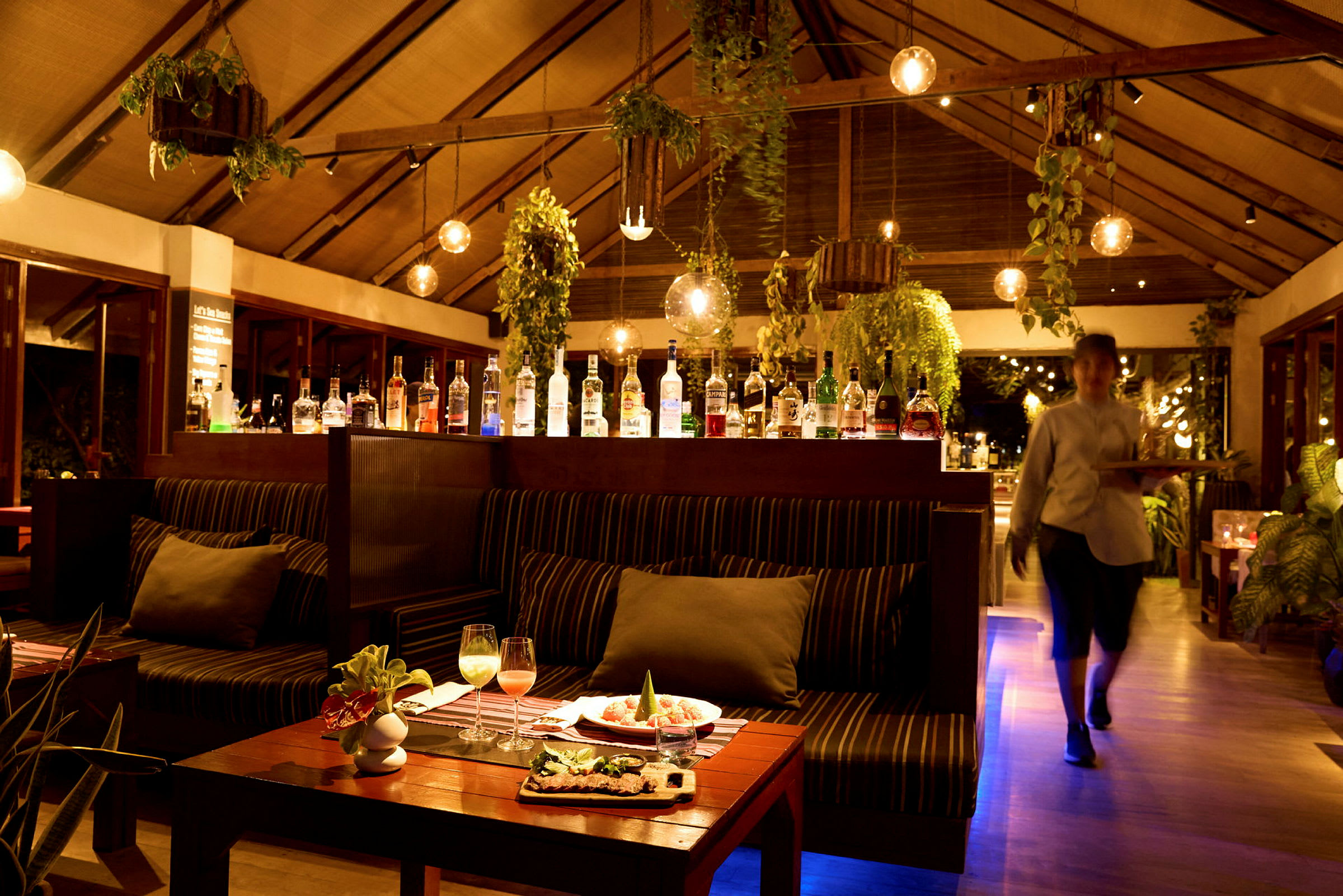 Interiør av en restaurant med sitteplasser, hengende planter og en bar som viser frem forskjellige spritflasker. (AI-generert tekst)