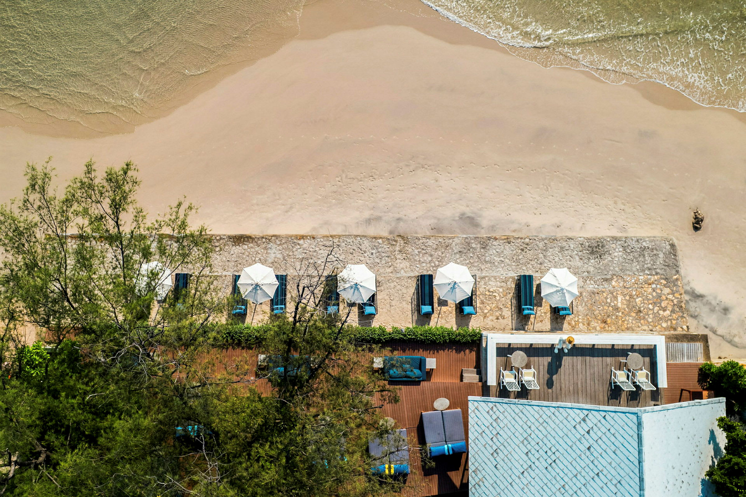 Utsikt ovenfra av en sandstrand med parasoller og solsenger samt en nærliggende bygning med terrasse. (AI-generert tekst)