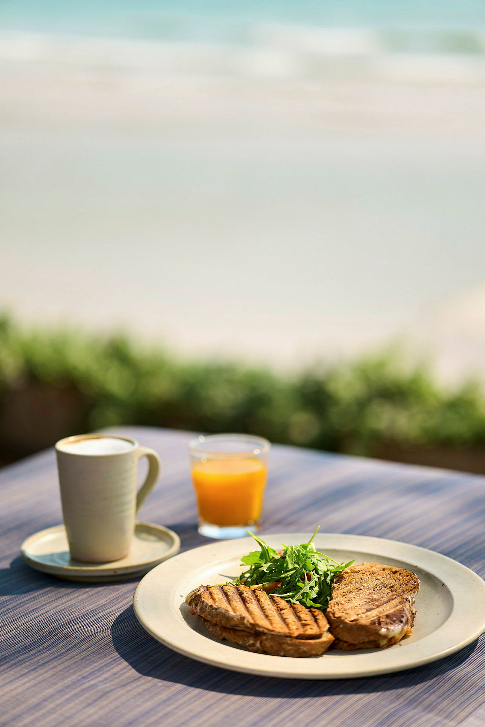 En tallerken med grillede smørbrød og salat, et glass appelsinjuice og en kopp kaffe står på et bord ved stranden. (AI-generert tekst)