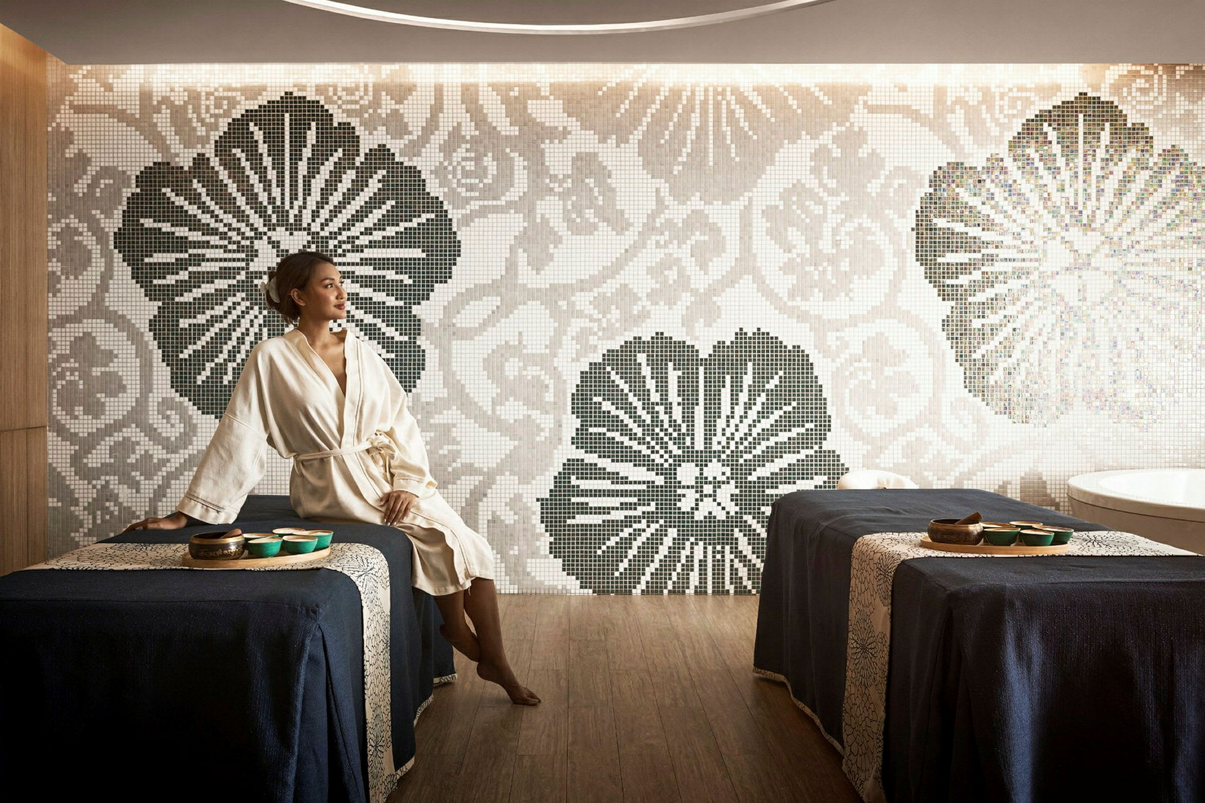 Et spa med to massasjebord dekket med mørkeblått stoff, en mosaikkvegg med blomstermønster og et badekar i bakgrunnen. (AI-generert tekst)