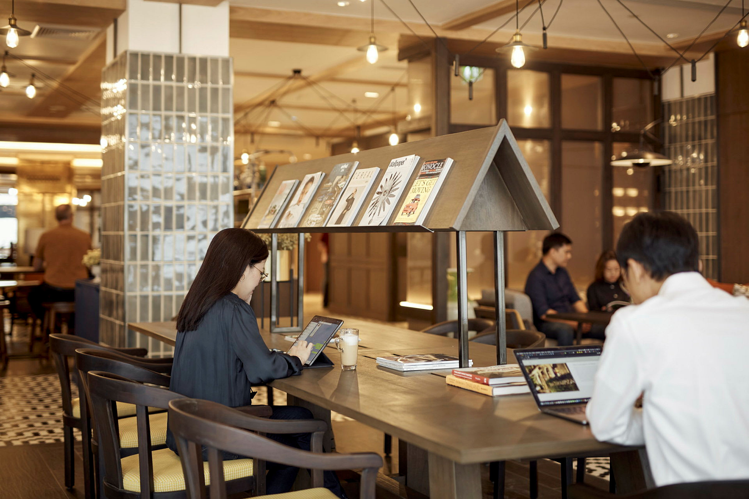 Et moderne kafé eller arbeidsområde med sitteplasser og bord. På bordet er det tidsskrifter og personer som jobber med bærbare datamaskiner. (AI-generert tekst)