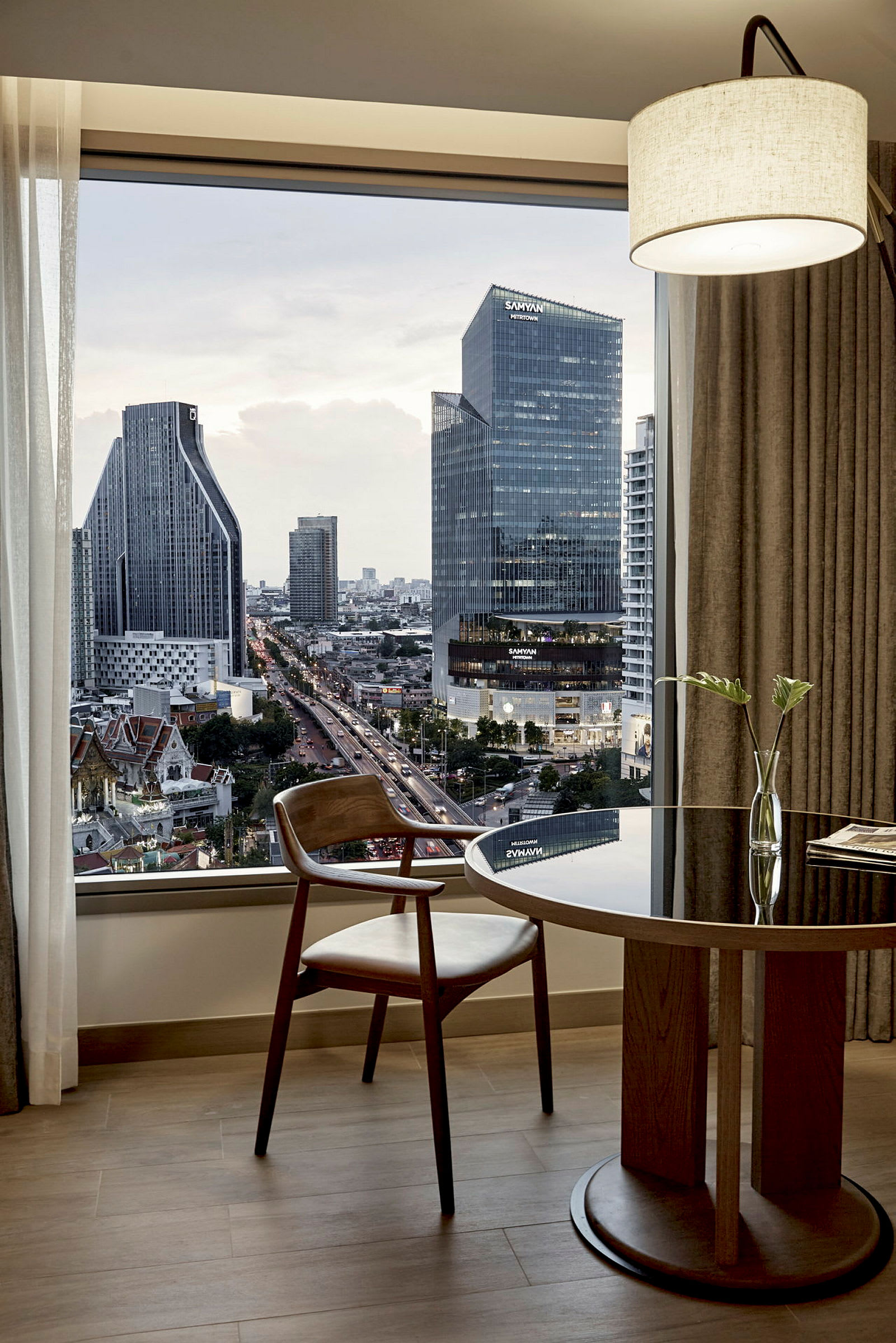 Et moderne hotellrom med utsikt over en by med høye bygninger i kveldslys. Et vindu viser utsikten, og foran det står et trebord med en stol og en enkel blomsterpotte. (AI-generert tekst)