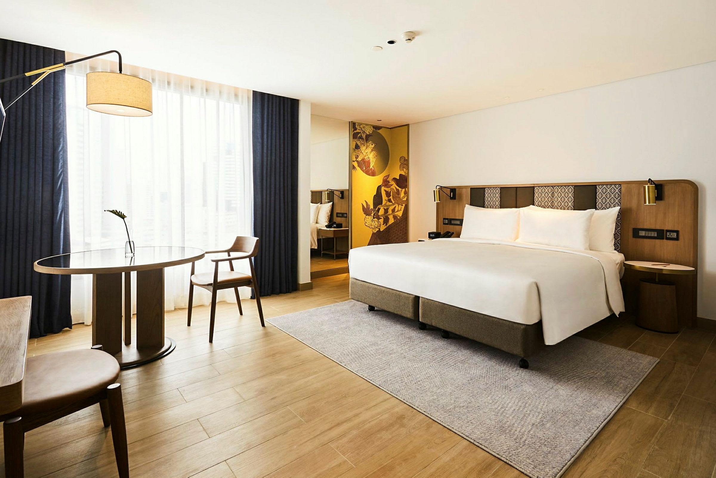 Et moderne hotellrom med en dobbeltseng, et bord og to stoler samt et stort vindusområde med gardiner. (AI-generert tekst)