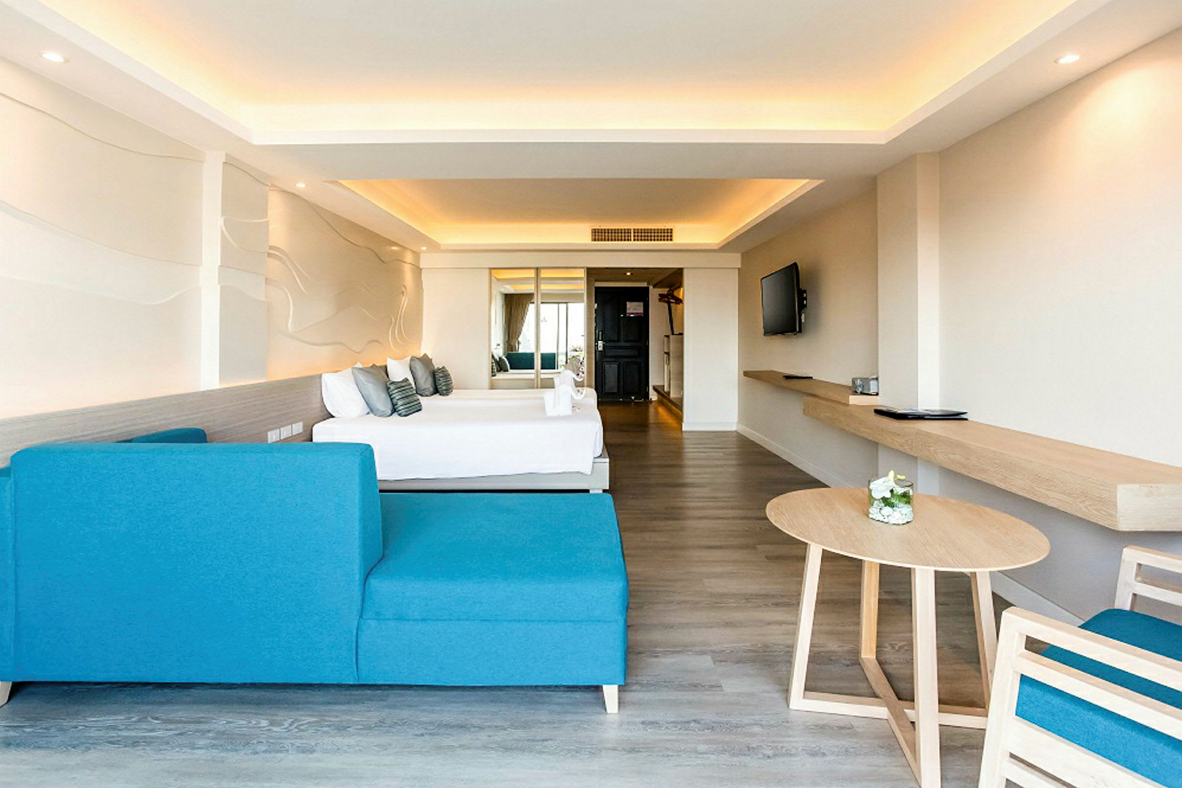 Et moderne hotellrom med en blå sofa, et bord og en seng i bakgrunnen. Veggen har en dekorativ design, og rommet er lyst med nøytrale farger. (AI-generert tekst)