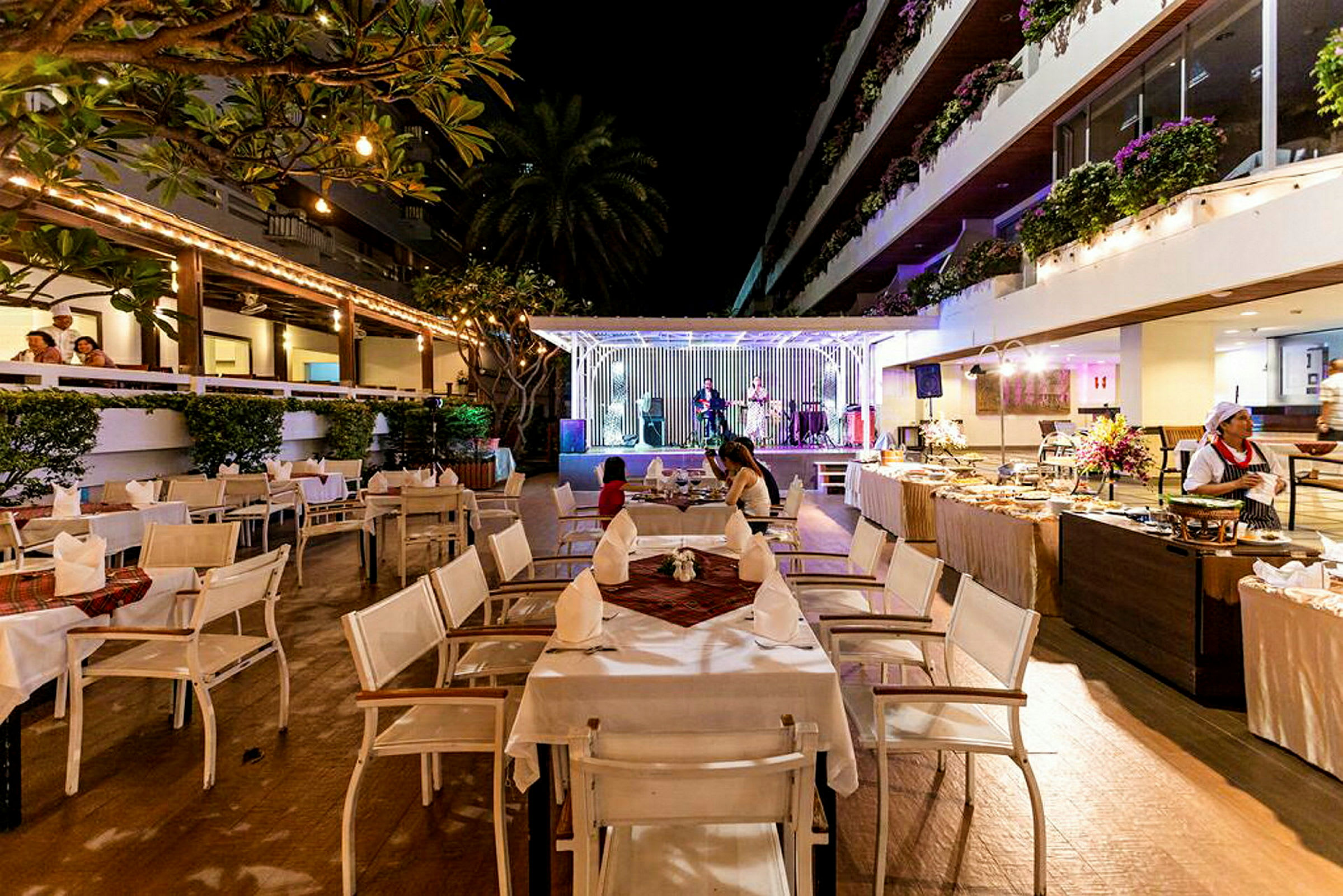 Bilde av en restaurant med uteservering om kvelden. Bord og stoler er oppstilt, og i bakgrunnen er en forhøyet scene med musikere. (AI-generert tekst)
