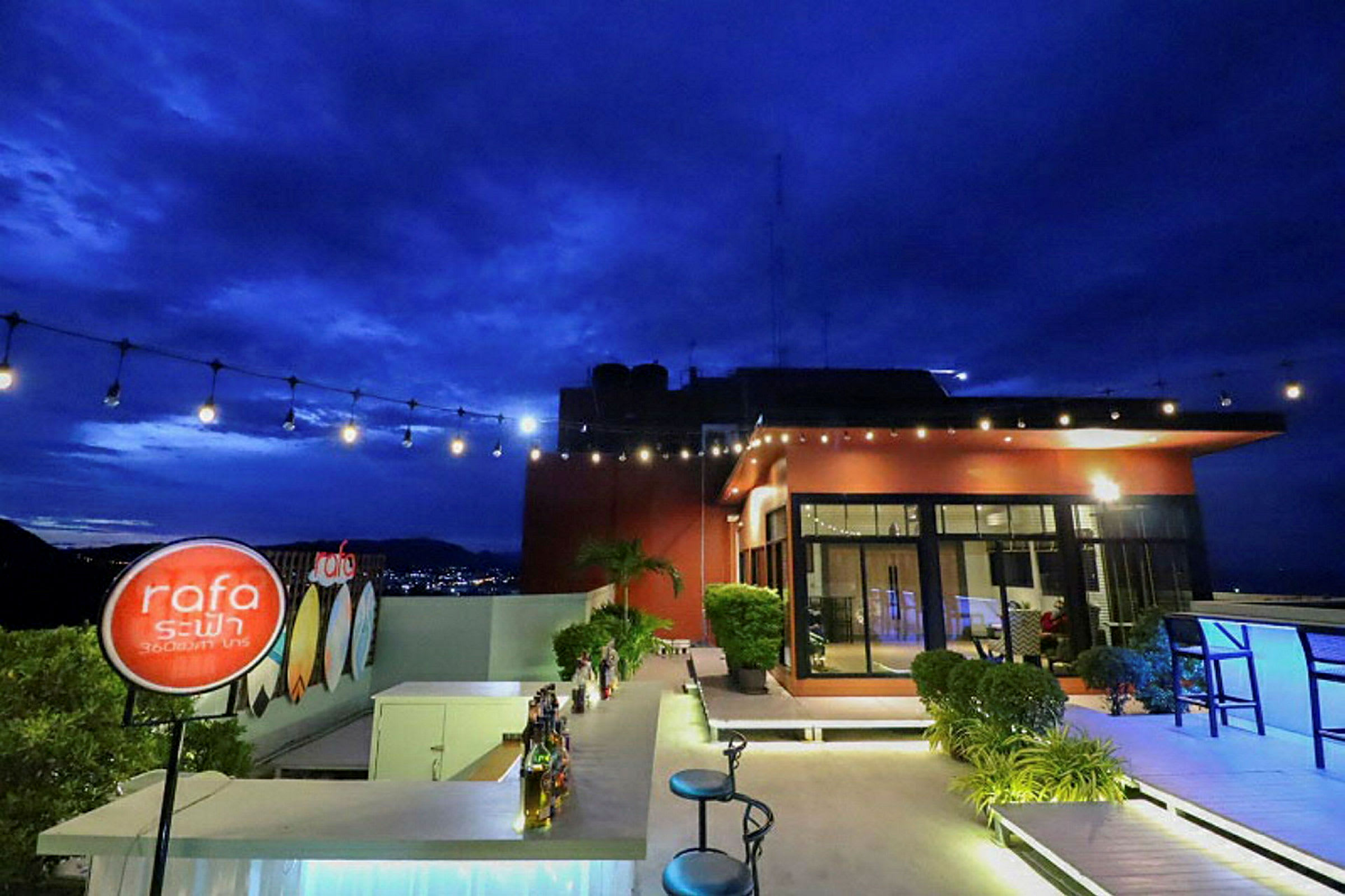 En takterrasse med en bar og stoler. Lys henger over, og i bakgrunnen ses en bygning og en by med nattehimmel. (AI-generert tekst)