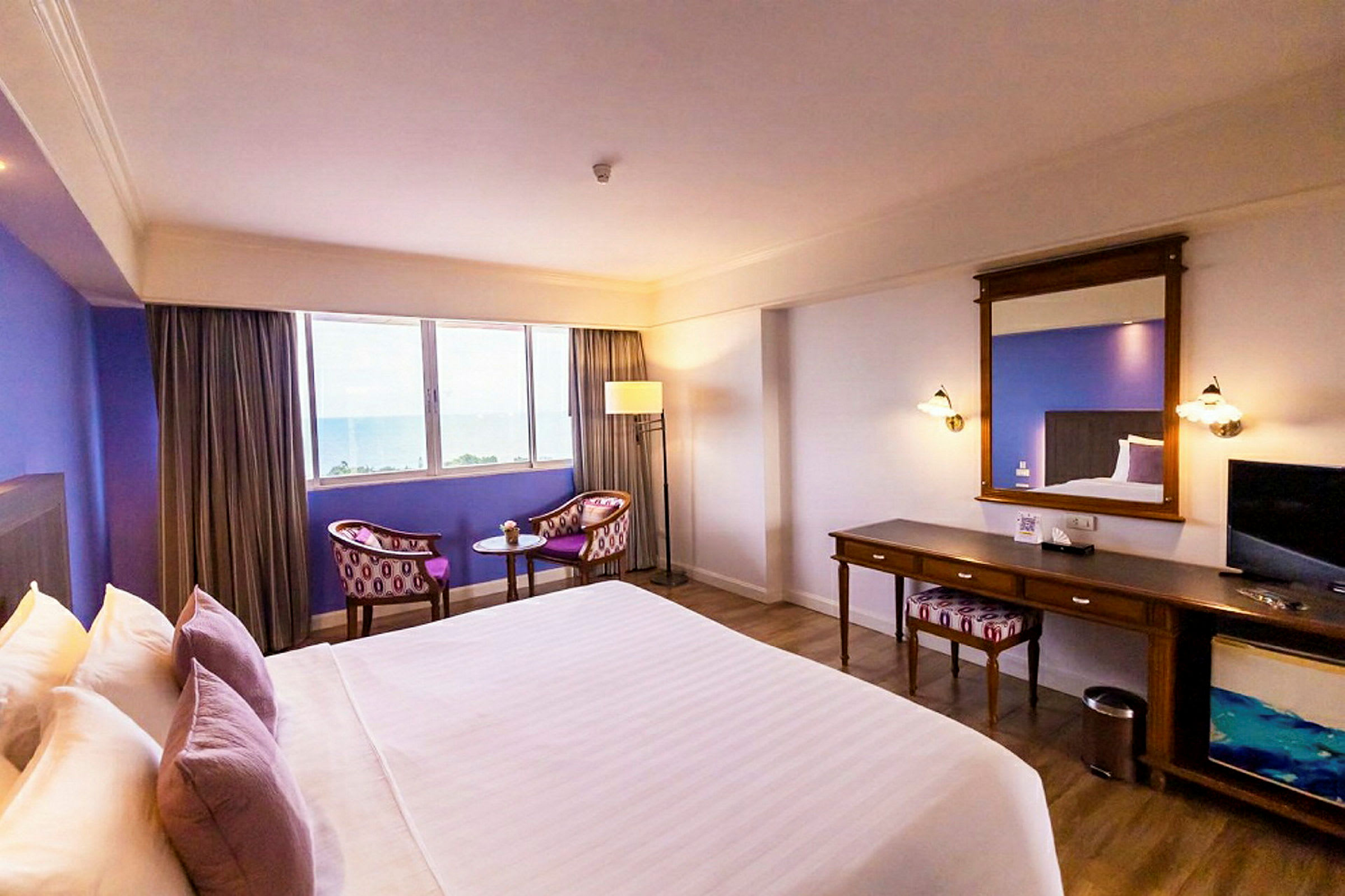 Et hotellrom med en stor seng, sitteområde ved et vindu og et skrivebord med speil. (AI-generert tekst)