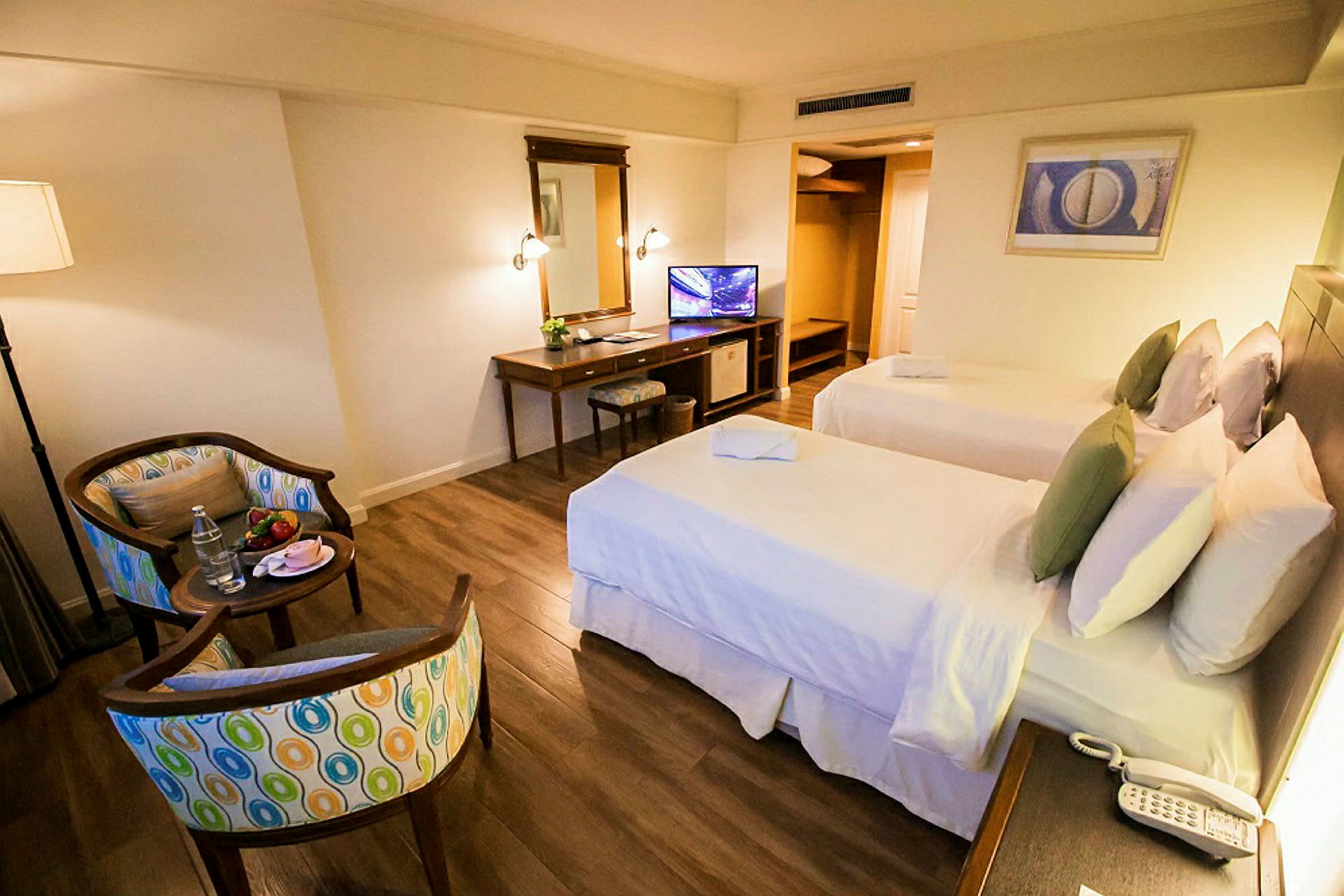 Et hotellrom med to separate senger, skrivebord, stol, speil og en TV. Rommet har tregulv og en sittegruppe med to stoler og et lite bord. (AI-generert tekst)