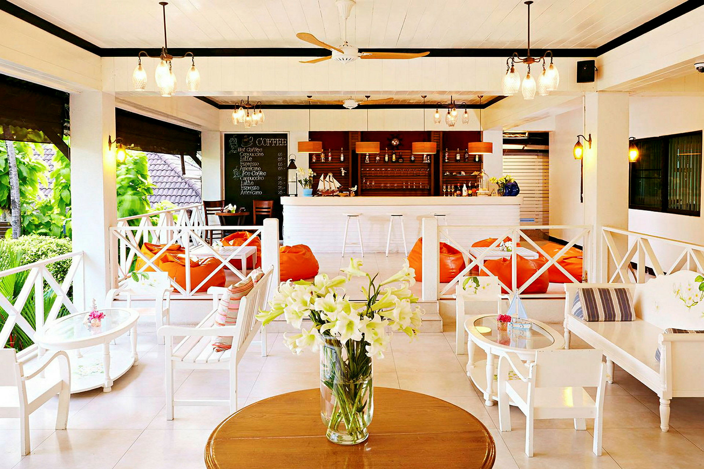 En lys og åpen lounge med hvite møbler, oransje sittesekker og en bar med drikkevarer i bakgrunnen, samt en vase med hvite blomster på et bord. (AI-generert tekst)