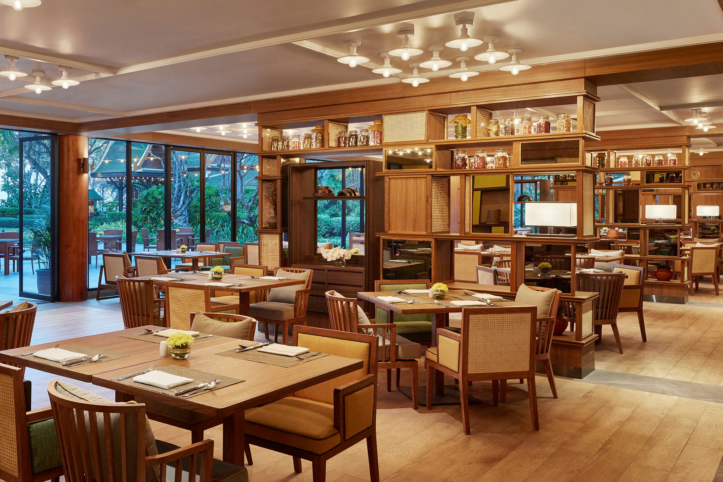 En elegant restaurant med tremøbler, bord og stoler arrangert i en åpen sal. Store vinduer gir utsikt mot grønne områder utendørs. (AI-generert tekst)