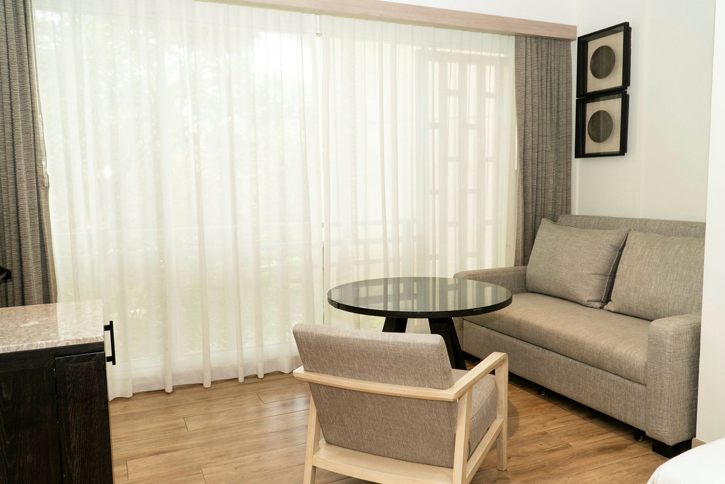 Et rom med en sofa og en stol i grått, et svart glassbord og et stort vindu med hvite gardiner. (AI-generert tekst)