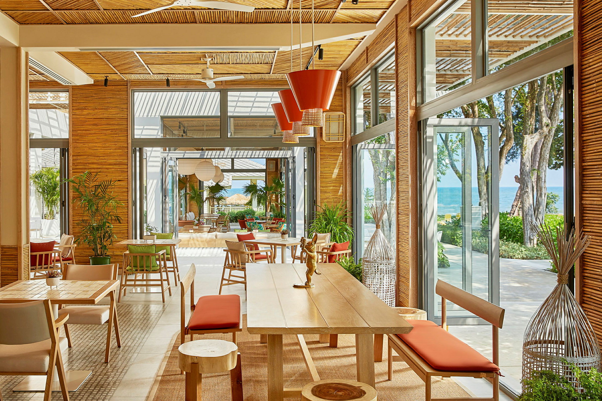 En lyst og romslig restaurant med store vinduer og utsikt mot havet. Møblene er moderne med tremateriale og røde detaljer. (AI-generert tekst)