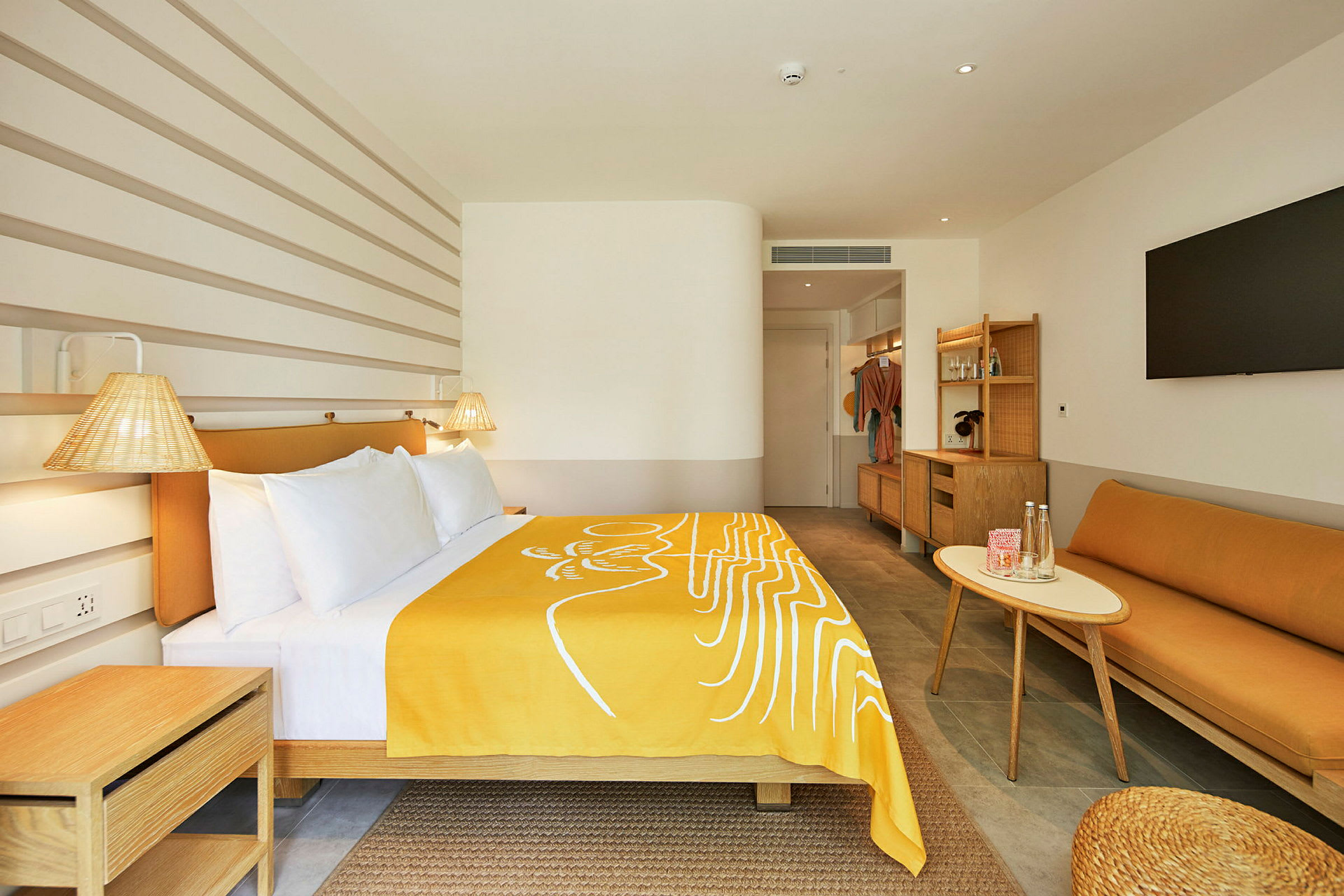 Et moderne hotellrom med en seng dekket av et gult teppe, en sofa, et bord og en stor TV på veggen. (AI-generert tekst)