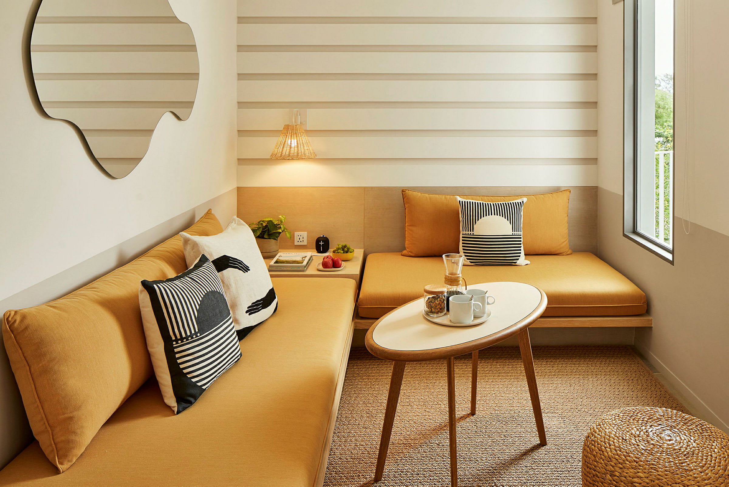 Et lite og moderne rom med to gule sofaer ved et lite, hvitt, ovalt bord. Veggene er hvite med horisontale striper, og et stort speil henger ved siden av sofaen. Vinduet til høyre slipper inn naturlig lys. (AI-generert tekst)