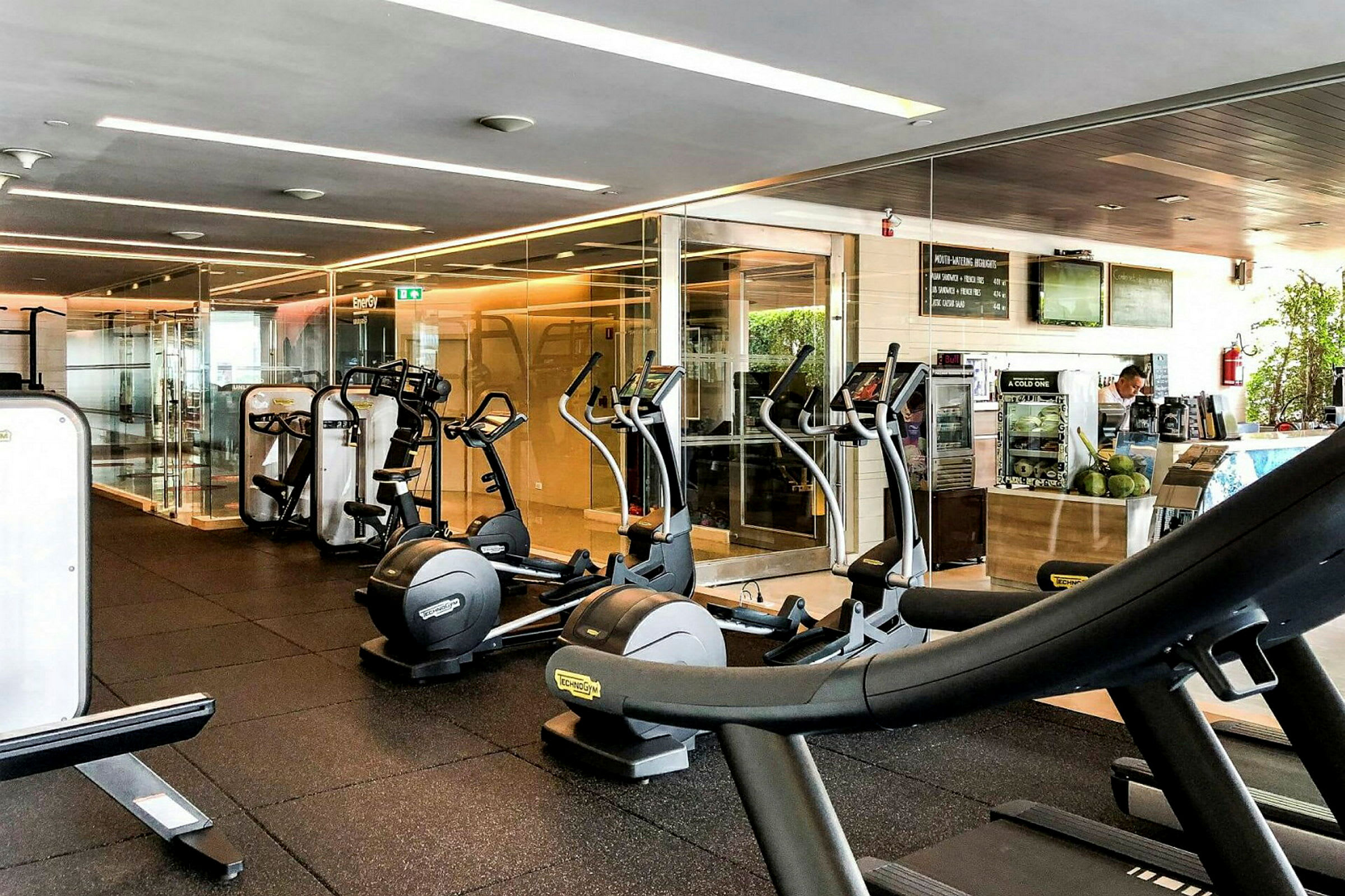 Et treningssenter med treningsutstyr som tredemøller og crosstrainere, samt en liten bardisk i bakgrunnen. (AI-generert tekst)