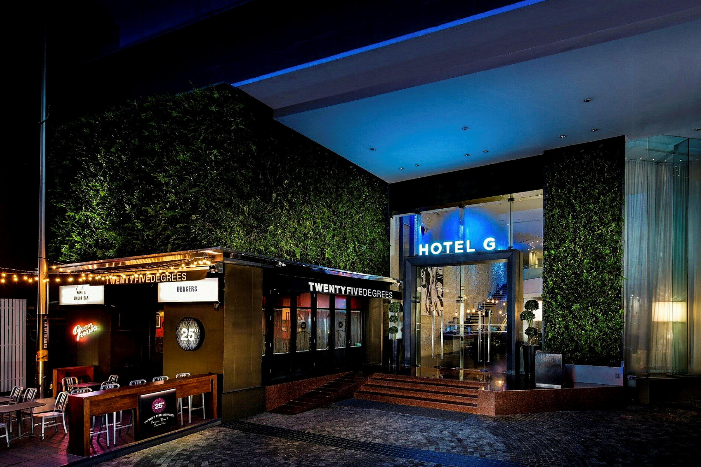 En opplyst hotellingang med en restaurant ved siden av, omgitt av grøntområder og moderne detaljer. (AI-generert tekst)