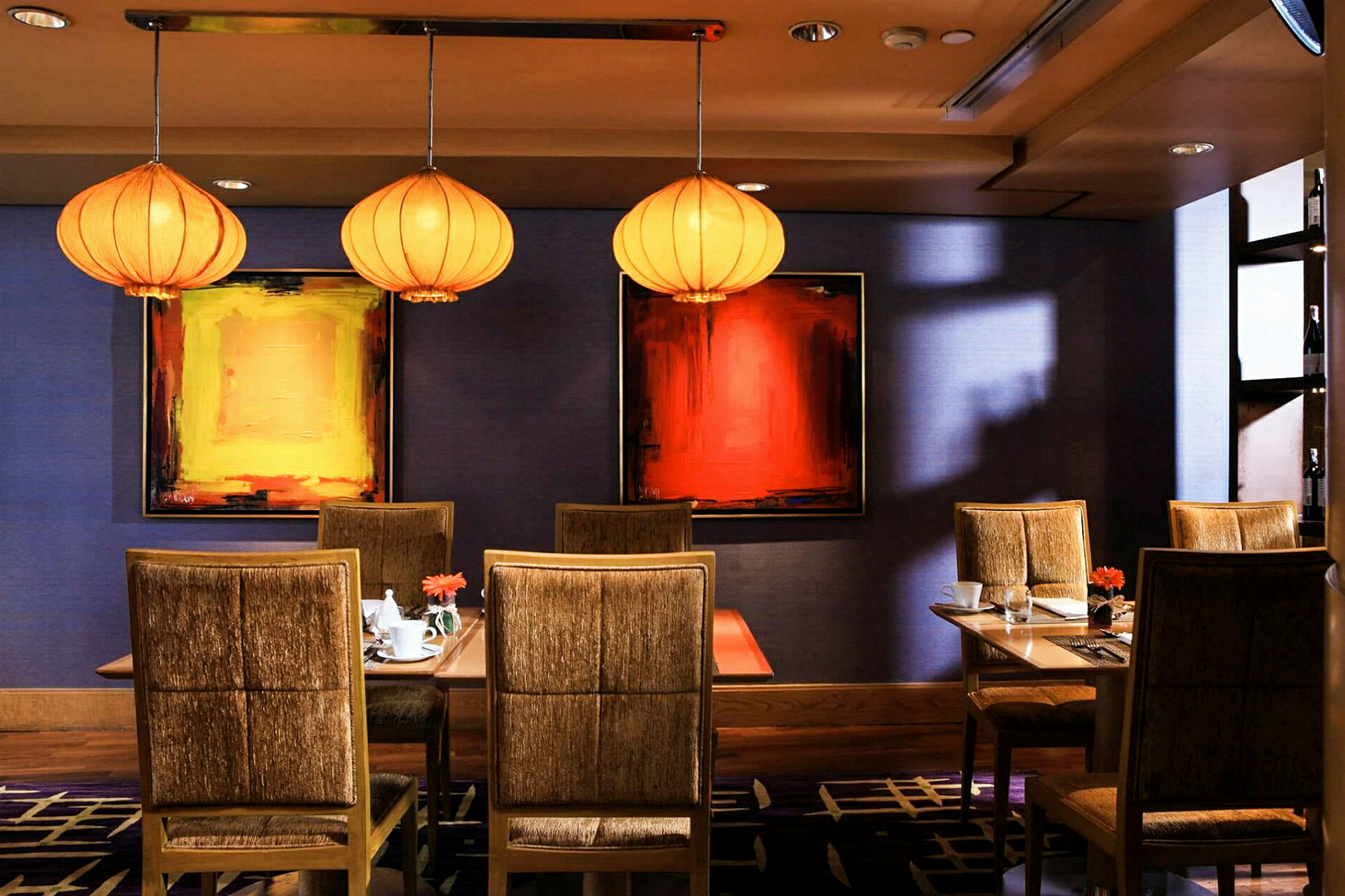 Et interiørbilde av en restaurant med tre bord omgitt av polstrede stoler, samt kunst på veggene og dekorative lamper i taket. (AI-generert tekst)