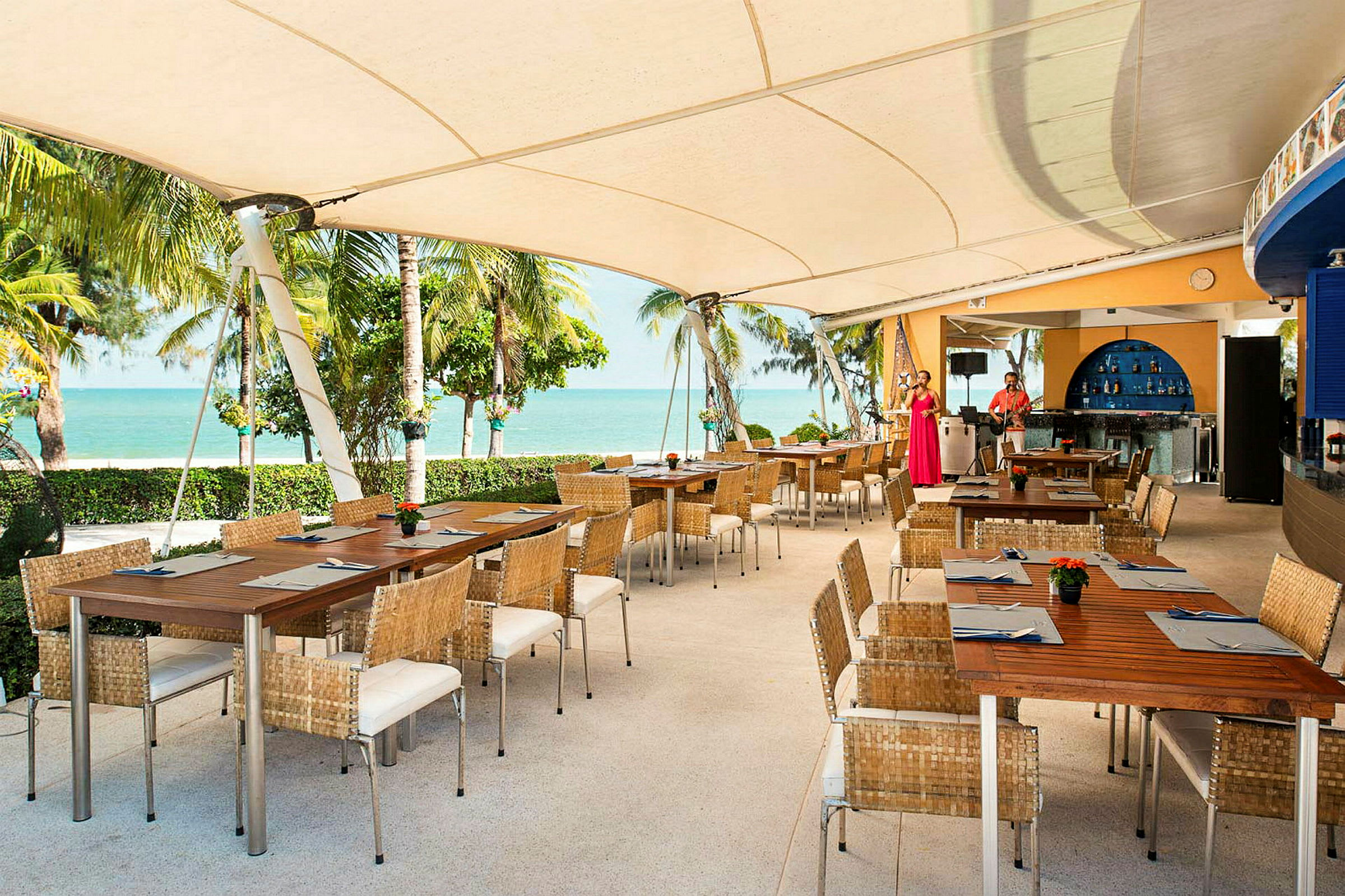 En utendørs restaurant med trebord og kurvstoler under et stort tak. Utsikt over stranden og havet med palmer i bakgrunnen. (AI-generert tekst)