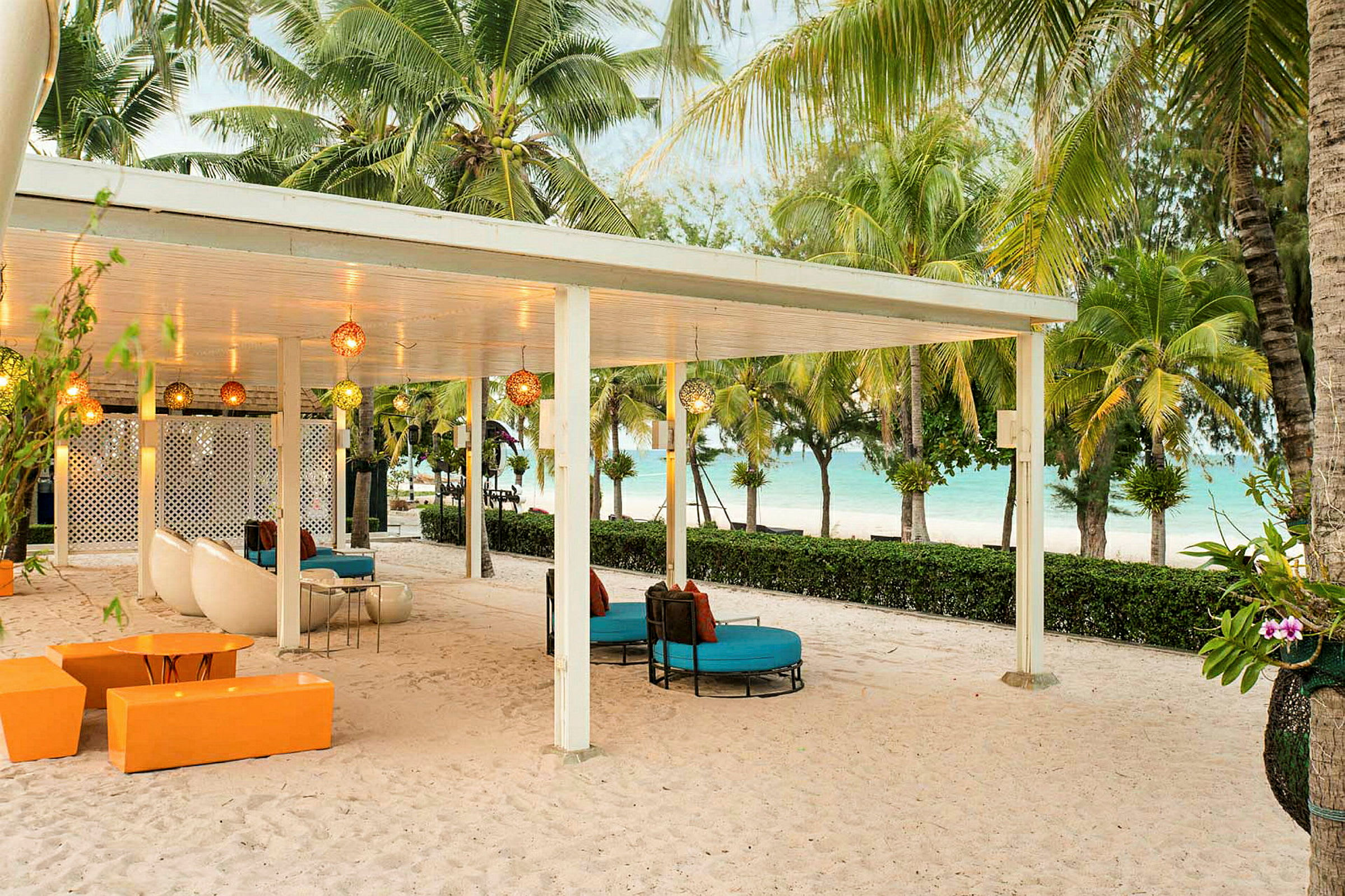 En overbygd uteplass med sittemøbler på en sandstrand, omgitt av palmer og med utsikt over havet. (AI-generert tekst)
