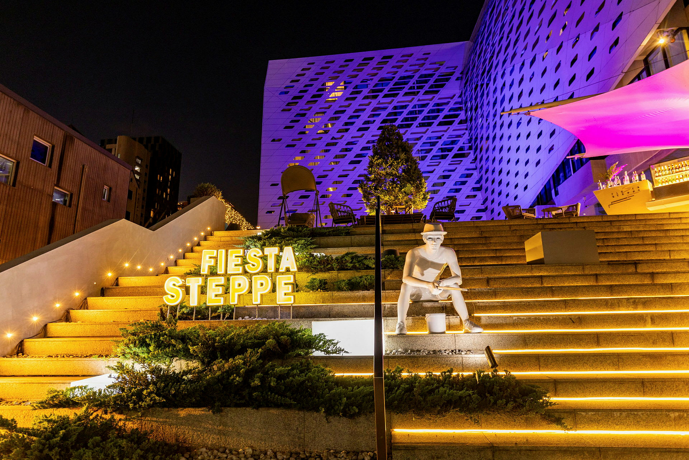 En utendørstrapp opplyst med gule lamper og dekorasjoner, med en statue og teksten 'Fiesta Steppe' synlig. (AI-generert tekst)