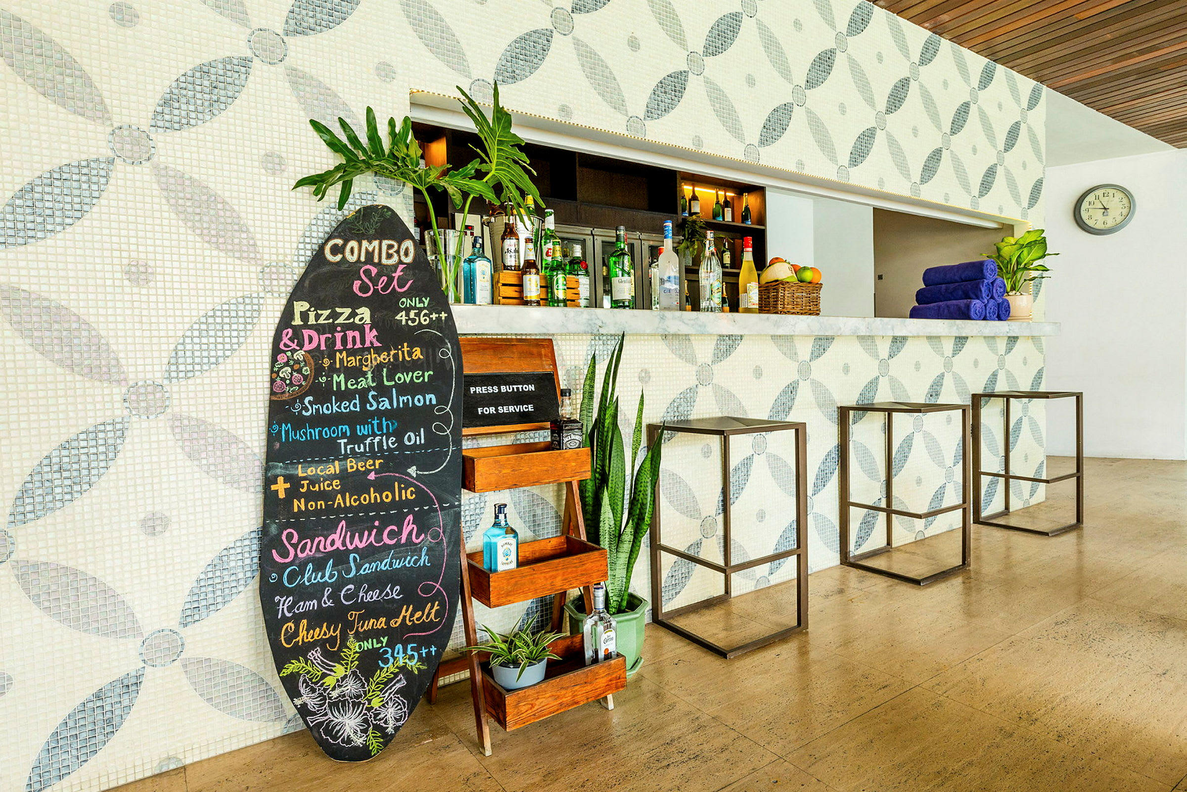Bildet viser en bar med flislagte vegger med mønster. Foran baren står et svart menybord med fargerik tekst, planter og dekorative gjenstander. Det er tre moderne barstoler og en klokke på veggen. (AI-generert tekst)