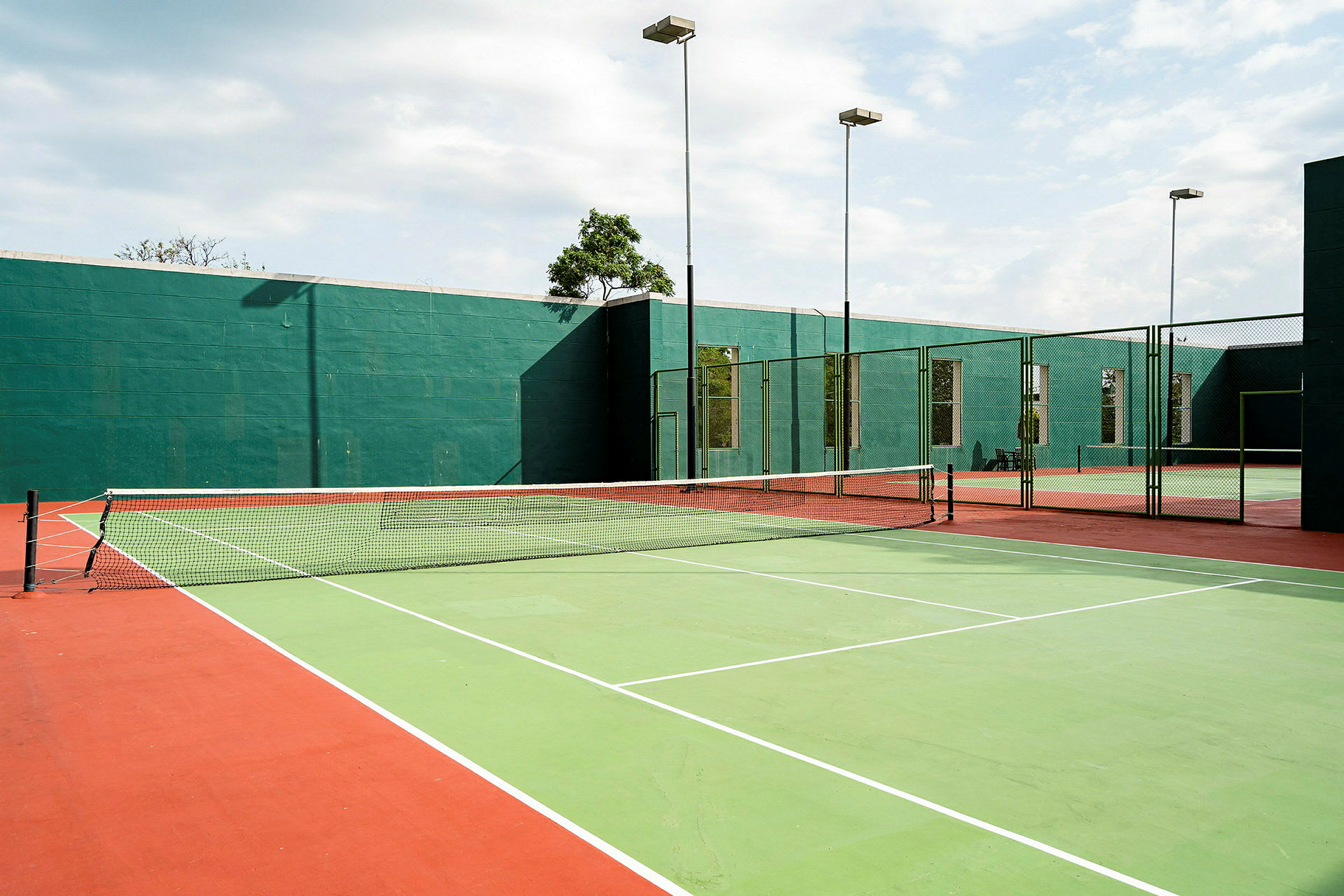 Utendørs tennisbaner med grønne og røde markeringer samt et nett i midten. Bak banene vises grønne vegger og høye stolper med lamper. (AI-generert tekst)