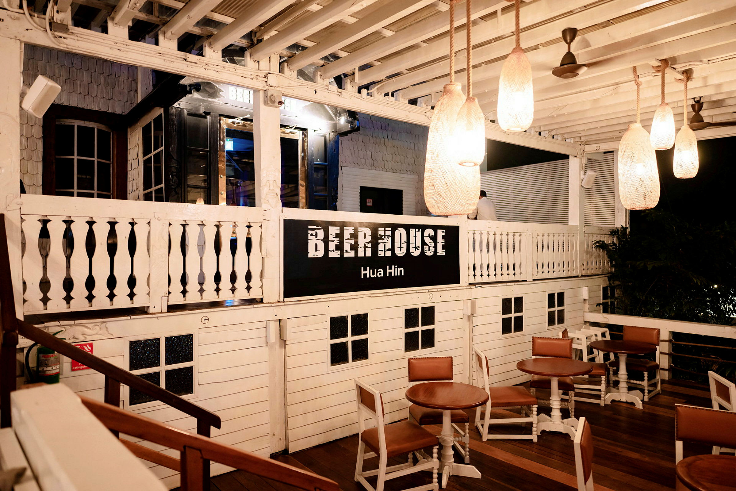 En trehus med skiltet 'Beer House Hua Hin'. Det finnes bord og stoler plassert på en utendørs veranda. (AI-generert tekst)