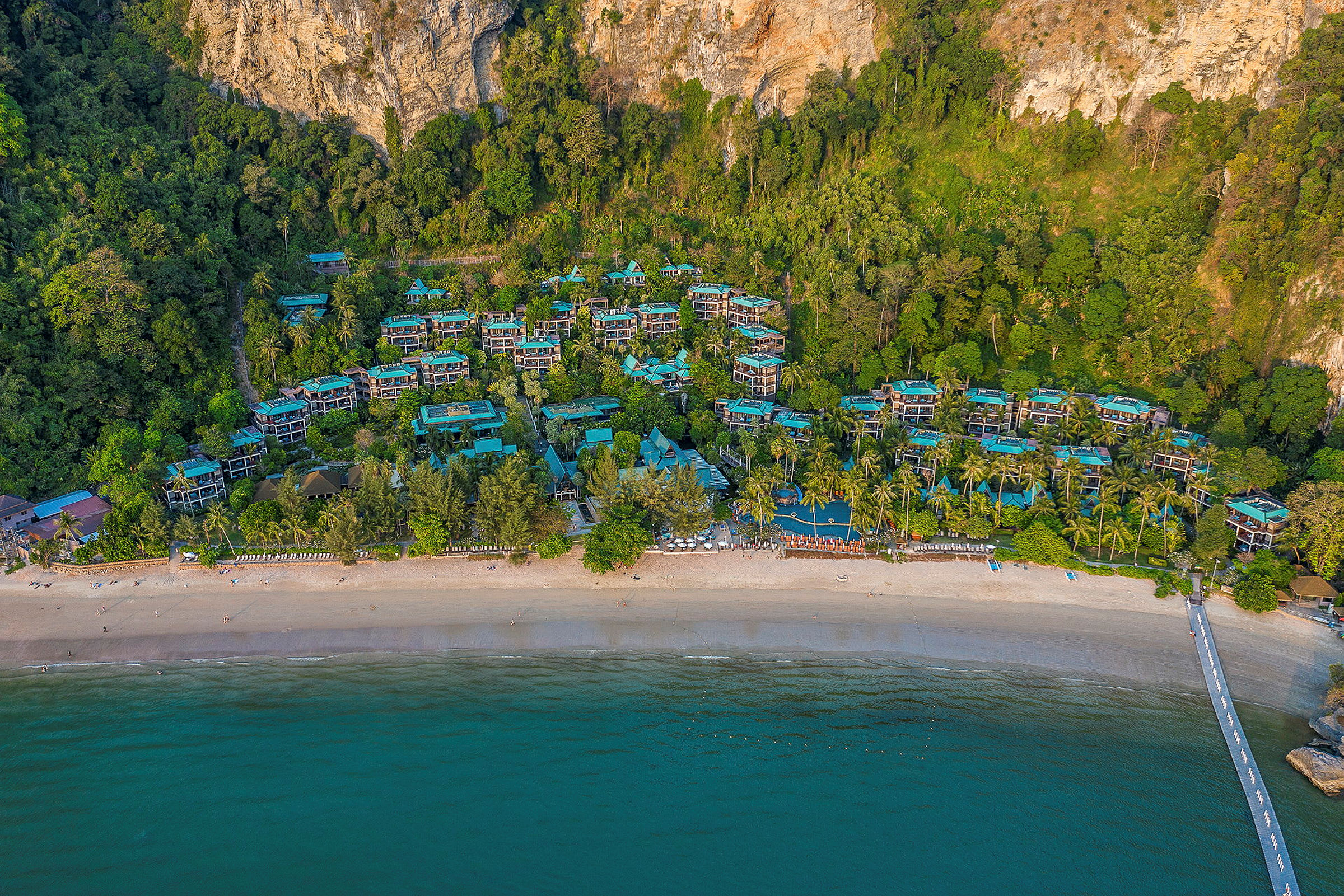 Luftfoto av en strand med turkisblått vann, hotellbygninger i naturskjønn beliggenhet og omkringliggende grøntområder. (AI-generert tekst)