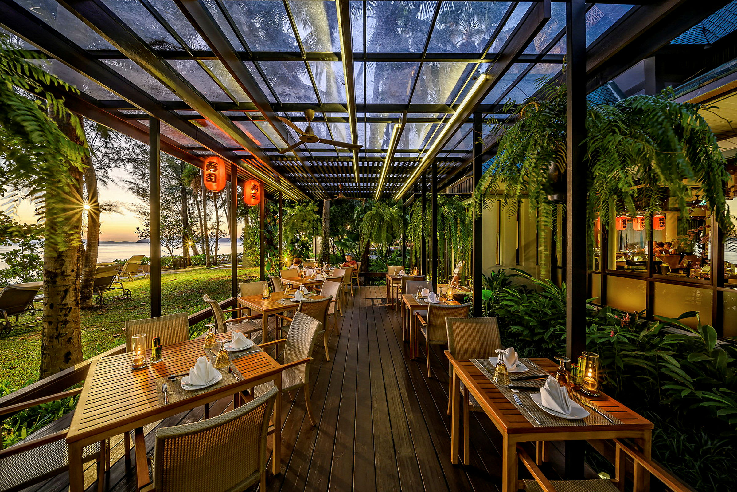 En restaurant med bord og stoler utendørs omgitt av mye grønt, med utsikt over en strand med solnedgangen i bakgrunnen. (AI-generert tekst)