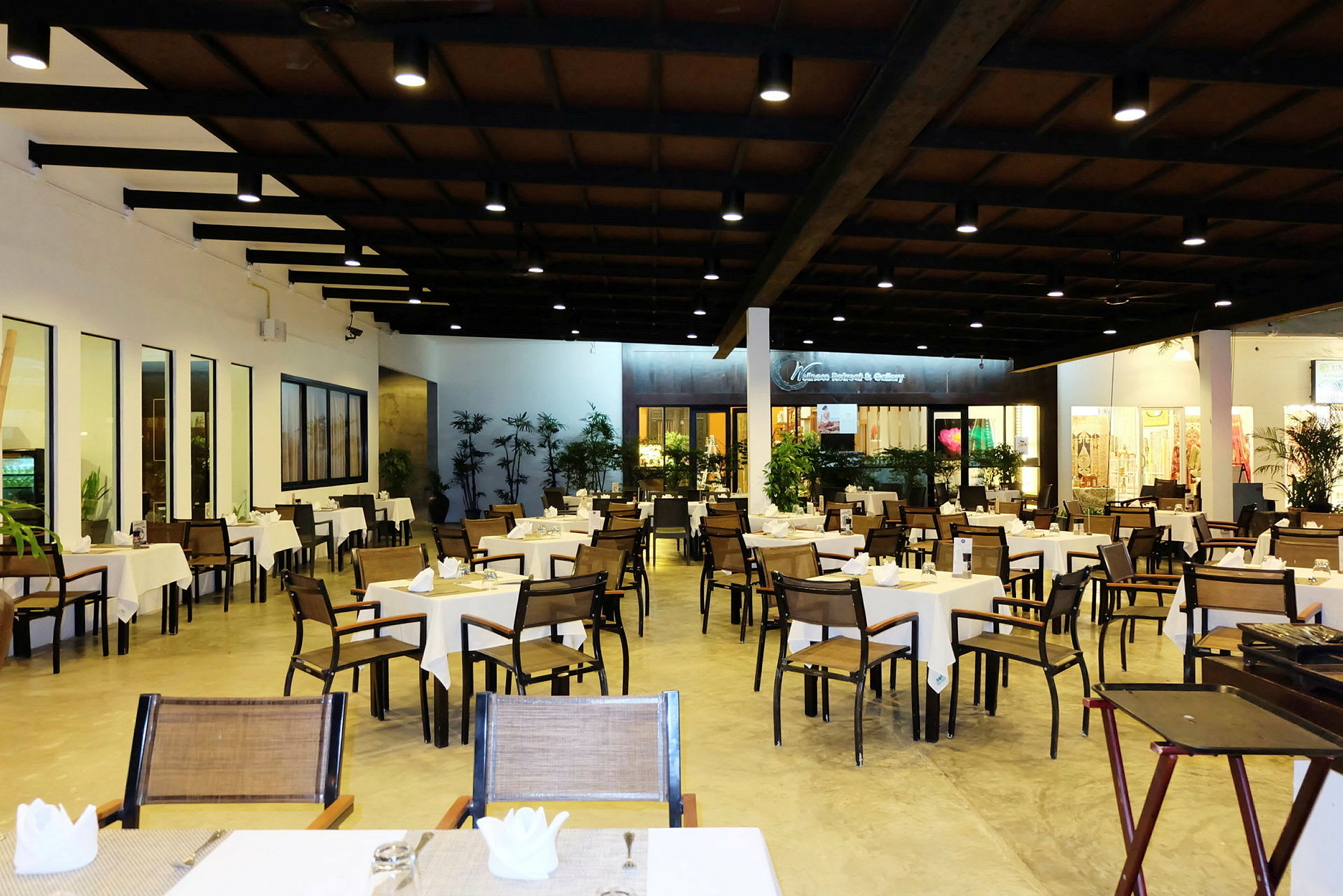 Et stort restaurantområde med mange bord og stoler, dekket for gjester, og omgitt av planter og lysbelysning. (AI-generert tekst)