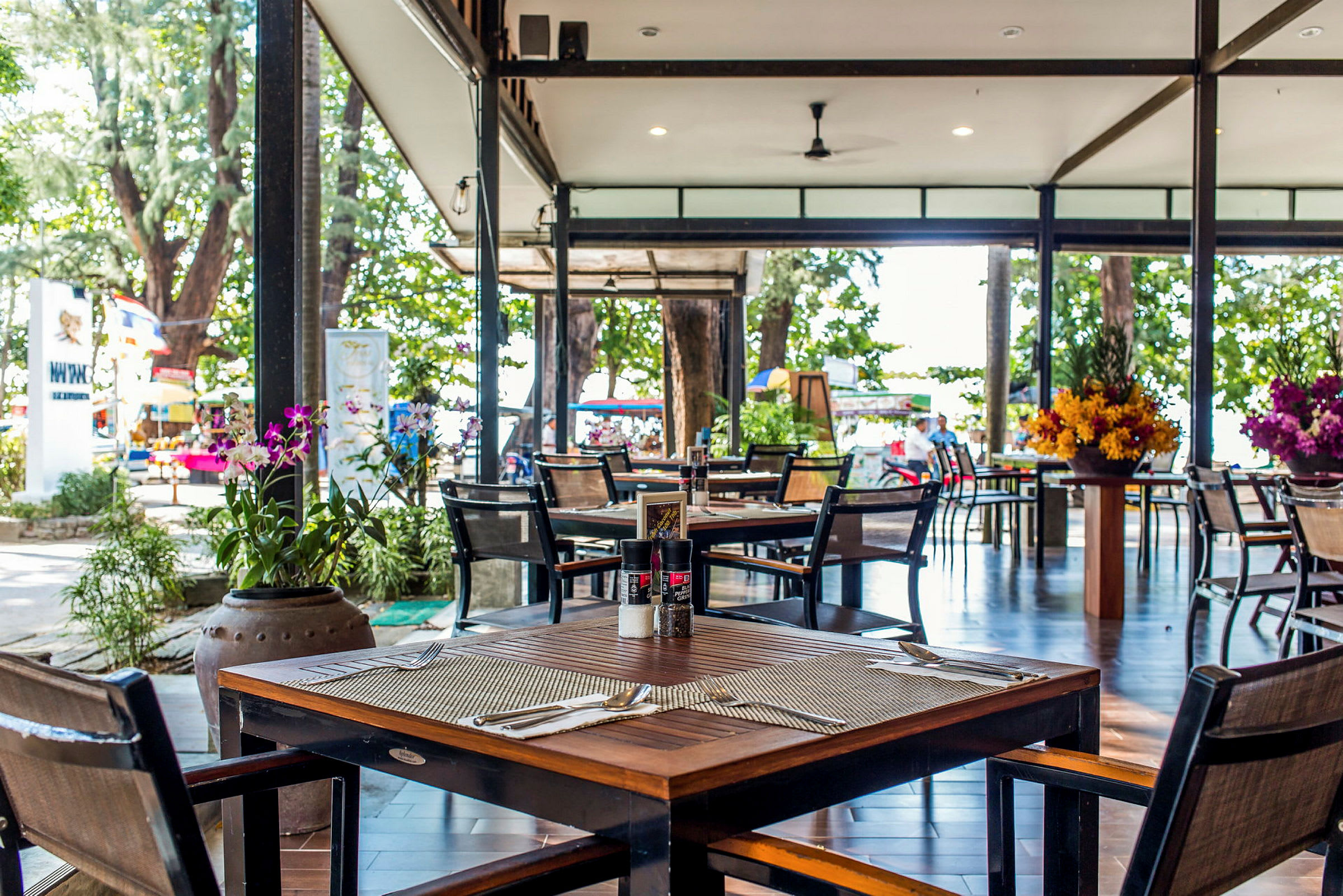 En utendørsrestaurant med møblerte bord og stoler. Store blomsteroppsatser pryder omgivelsene, og i bakgrunnen sees grønne trær og en strand. (AI-generert tekst)