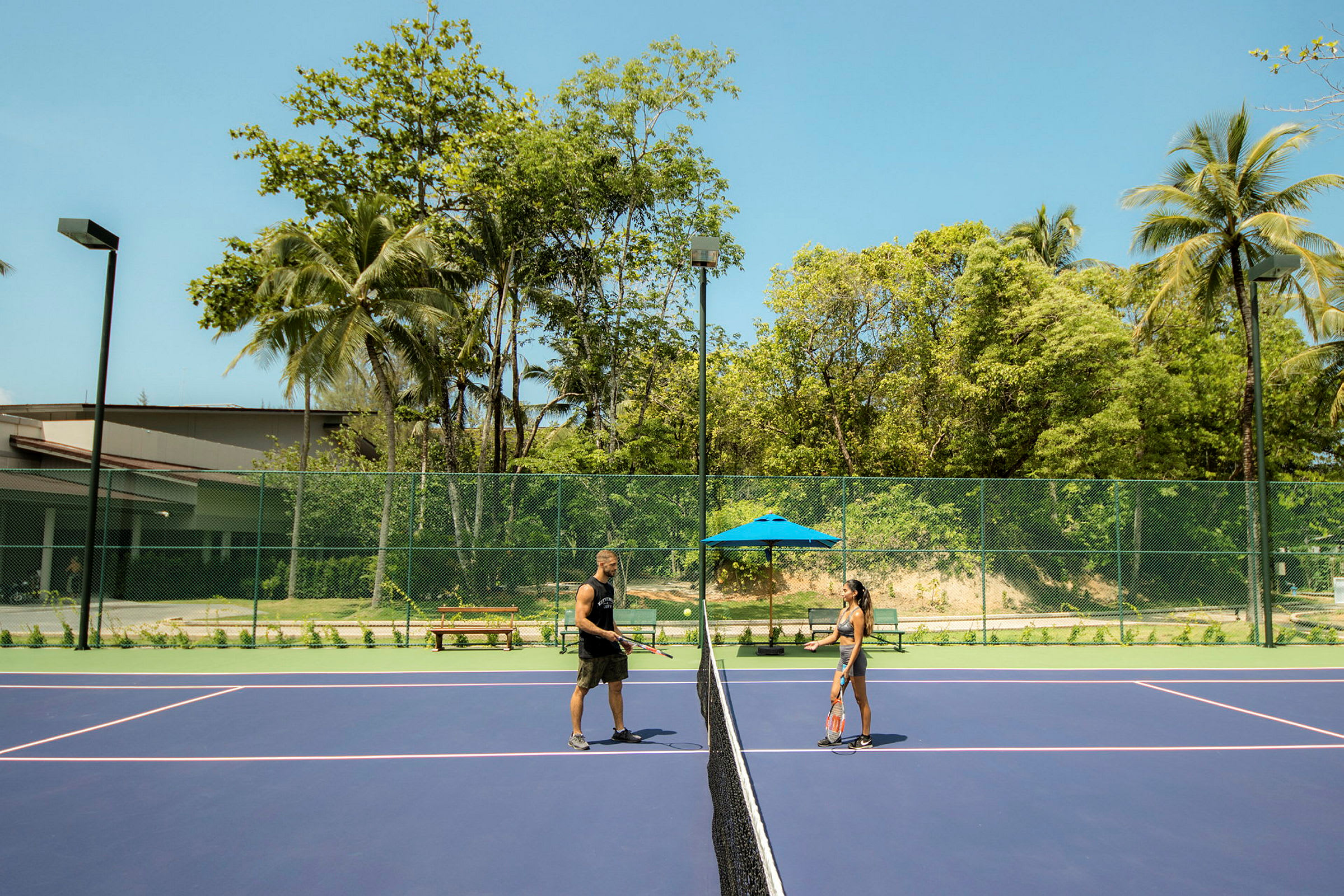 To personer spiller tennis på en utendørs bane omgitt av trær. (AI-generert tekst)