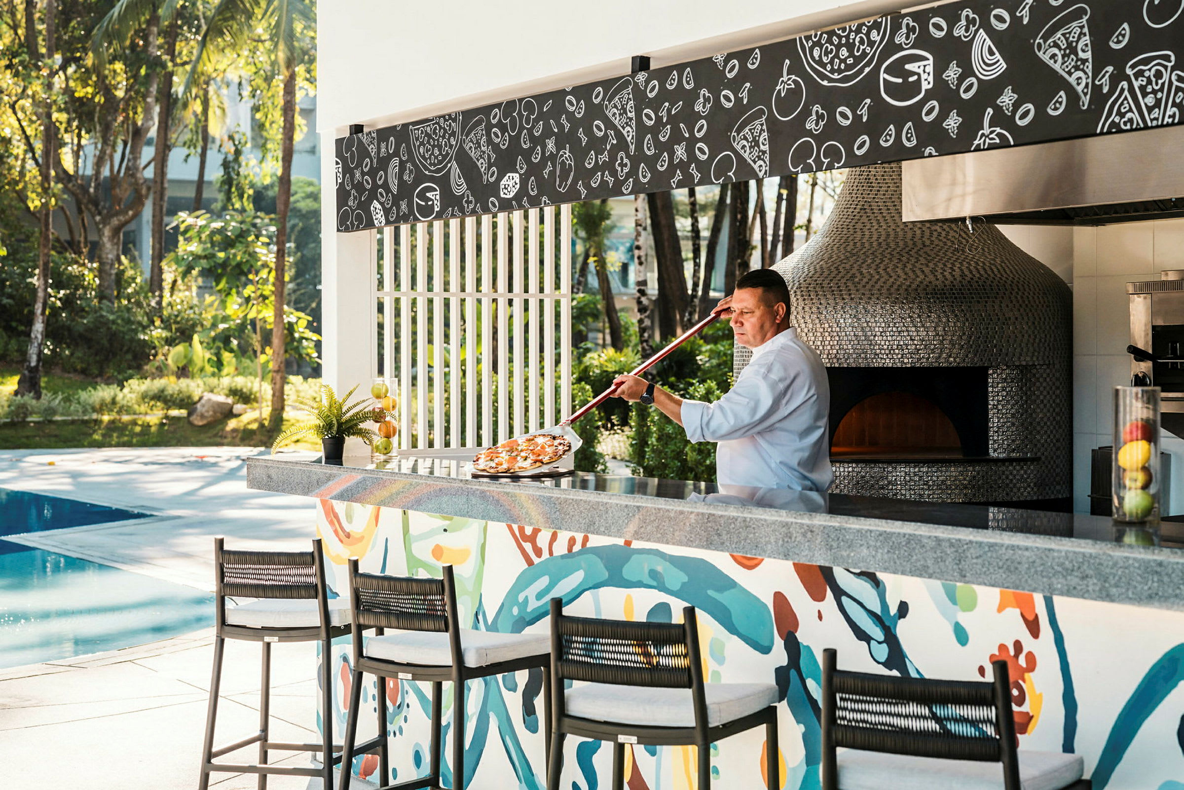 En pizzakokk plasserer en pizza i en vedfyrt ovn ved en utendørsbar nær et basseng. (AI-generert tekst)