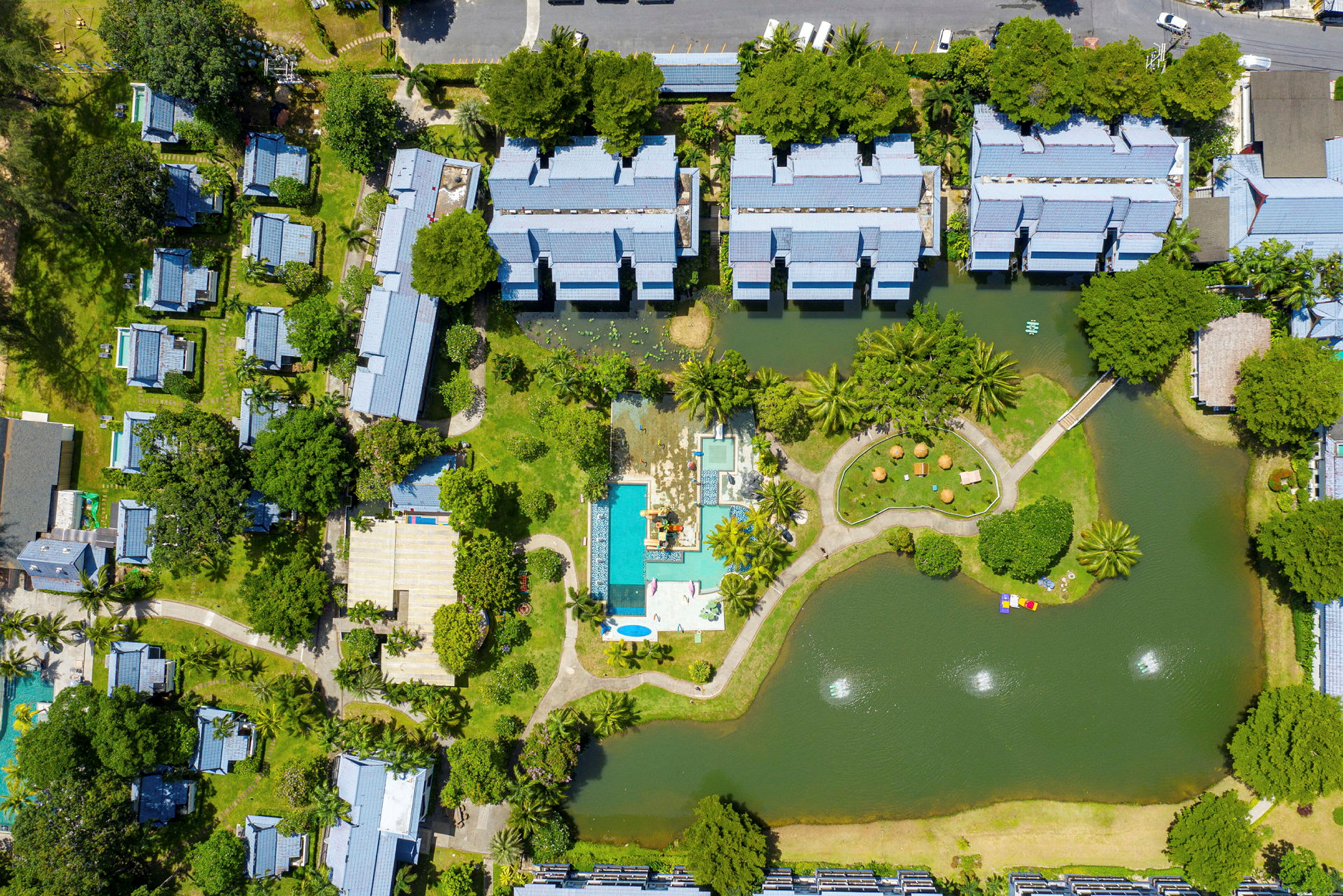 Flyfoto av en resort med bygninger, bassengområde og omkringliggende grøntområder. (AI-generert tekst)